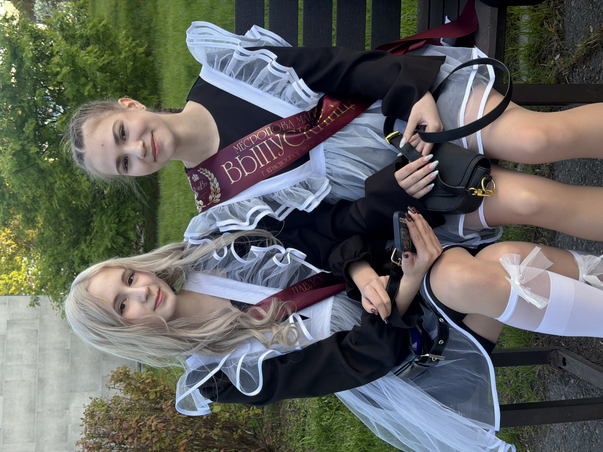 Two fair-skinned young women sit side-by-side on a dark bench in an outdoor setting with greenery in the background. Both wear similar black and white school uniforms with ruffled collars and graduation sashes bearing the inscription "ВЫПУСКНИЦ" (Graduate) in Russian. One sash bears the full name "МЕСРОПОВА МАРИЯ" (Mesropova Maria), and the city "Г. КРАСНОЯРСК" (Krasnoyarsk). The blond woman sits at the front of the image while the brunette is behind her. Both girls are holding black handbags. The background includes trees and grass. The location is the city of Krasnoyarsk in Russia.