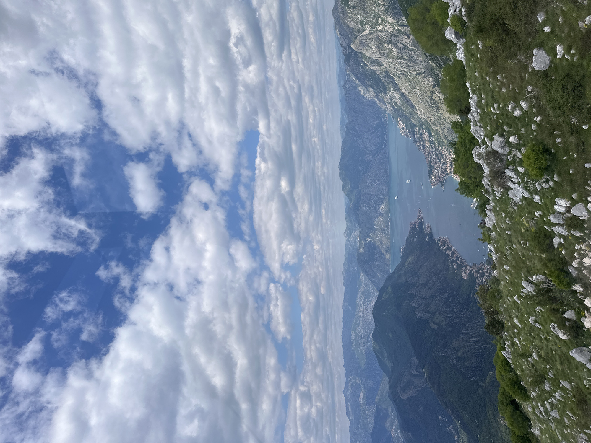 The image shows a mountain landscape with a body of water below and a cloudy sky above. In the foreground, there's a steep, rocky slope covered in vegetation. The water appears to be a lake or bay surrounded by mountains. Clouds fill a significant portion of the sky. The scene could be located near Škaljari, Montenegro, given the topography, but a definite confirmation would require specific landmarks.