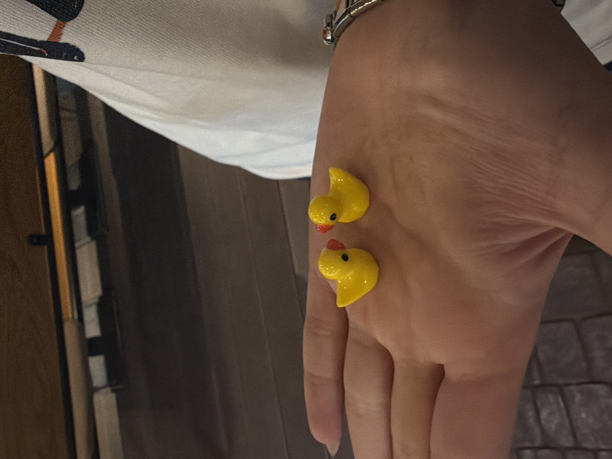 The image shows a person's hand with two small, yellow, plastic duck figures resting on the skin. The hand is positioned with the palm facing the camera. A bracelet is visible on the wrist. The background includes part of a white and black garment, a portion of a brown wooden structure, and part of a brick-paved area. There are no clear indications of it being in Yerevan, Armenia.