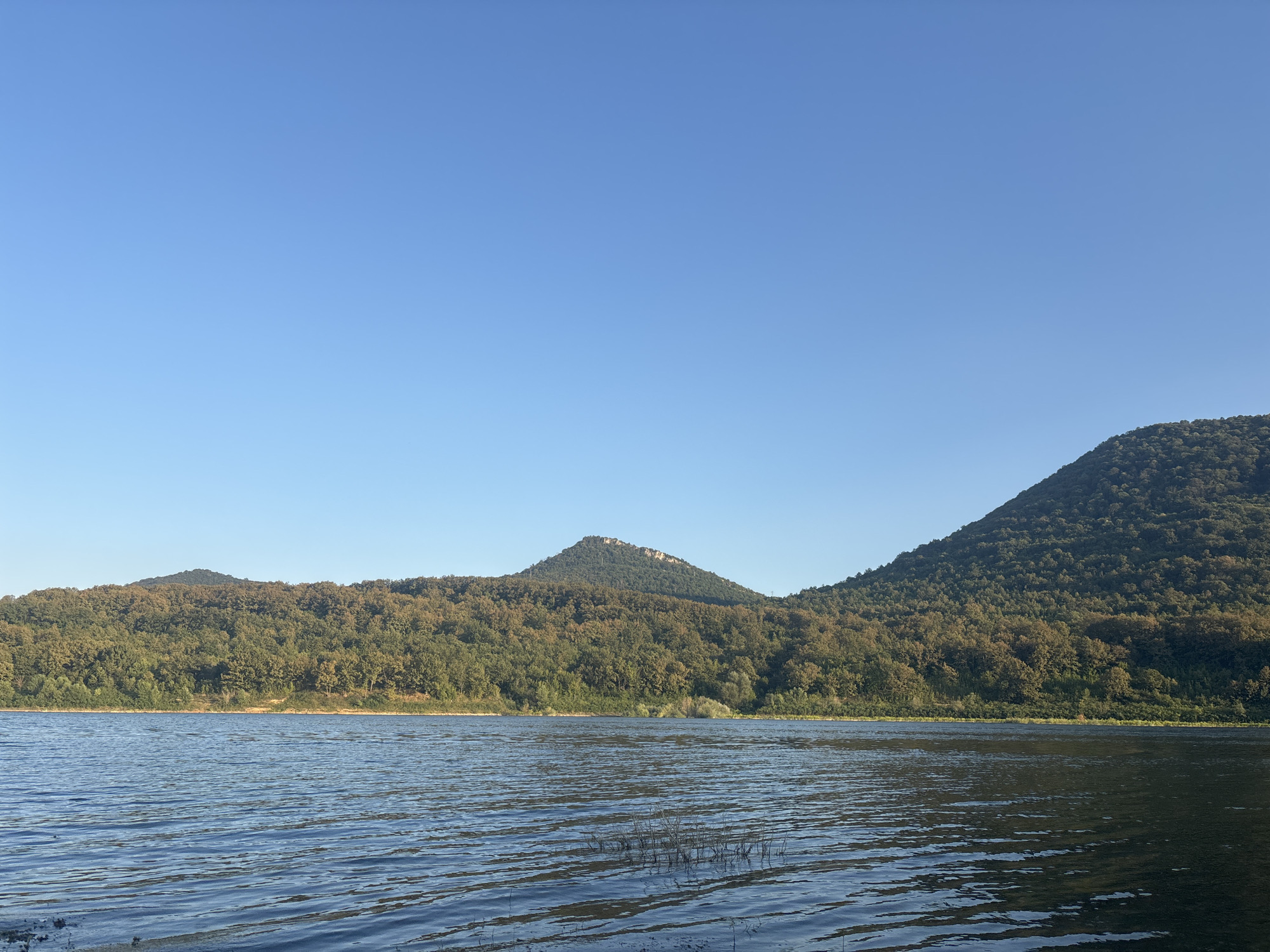 The image depicts a serene landscape with a clear blue sky dominating the upper portion. In the mid-ground, a dense forest covers rolling hills, with a prominent, cone-shaped peak in the center of the frame. The trees exhibit a mix of green and autumnal hues, suggesting it could be late summer or early fall. A body of calm blue water occupies the foreground, with subtle ripples on its surface. The scene is devoid of any human presence or activity, emphasizing a natural and tranquil environment. This landscape is consistent with the scenic beauty found around Asparuhovo, Bulgaria, known for its natural reserves and water bodies.