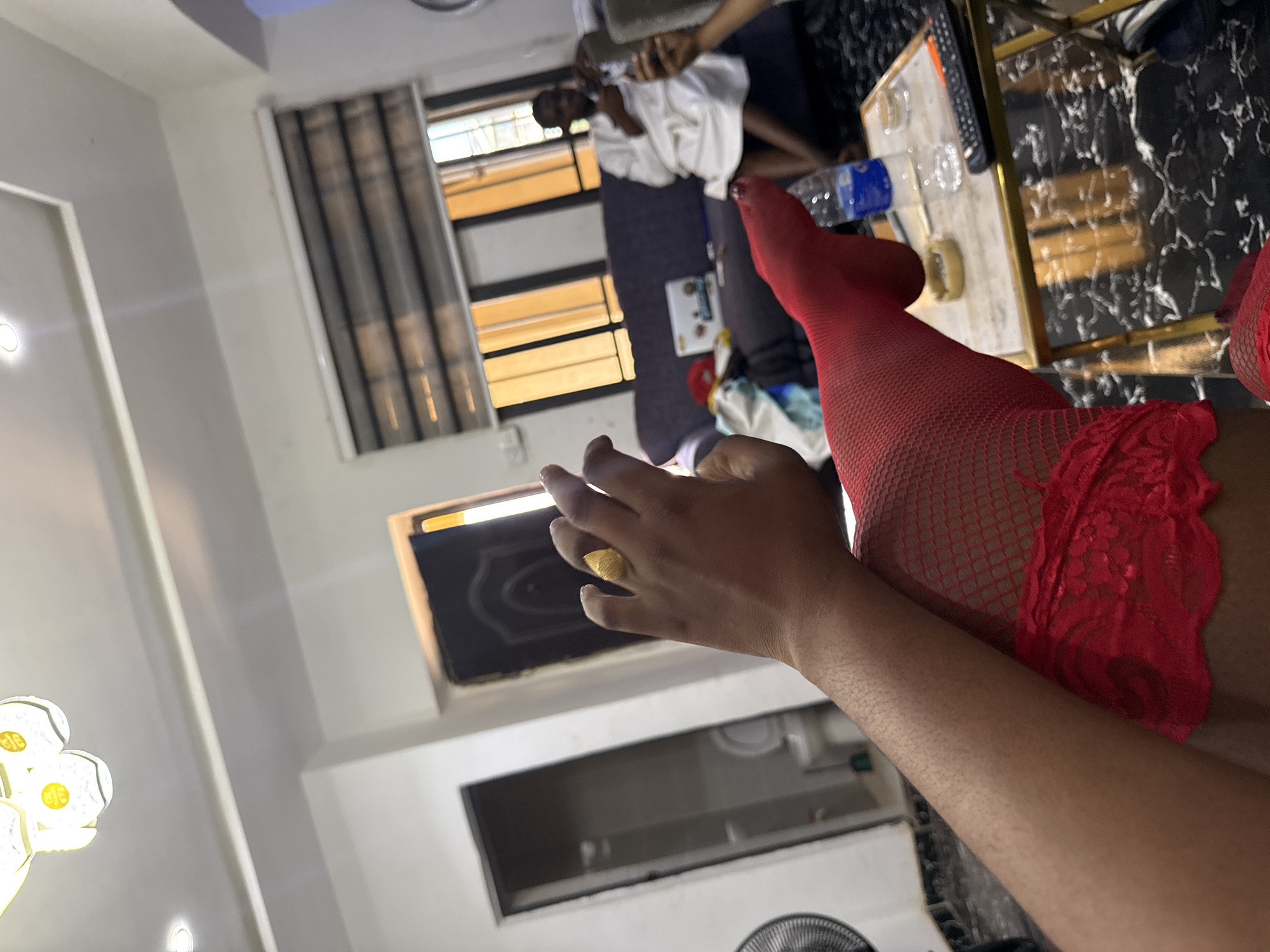 A photograph depicting a residential interior in Abuja, Nigeria. Focus on an individual wearing red fishnet stockings, seated near a black marble-topped table with a water bottle, glass, and remote control on it. In the background, a person wearing a white robe appears to be lying on a grey sofa. The room is lit by both natural light coming through window blinds and artificial lighting. A black door with intricate detailing is visible on one wall.