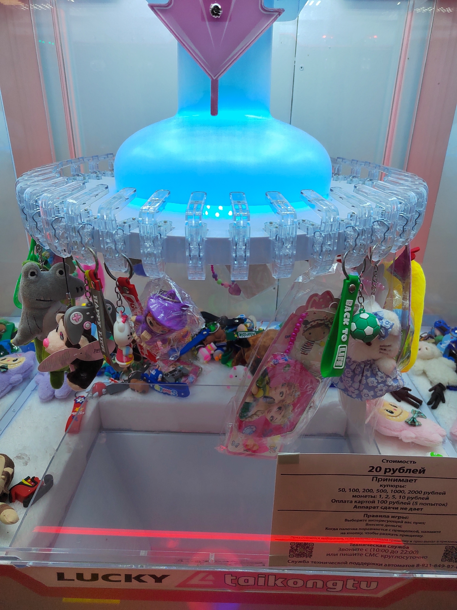 The image shows a claw machine filled with plush toys and keychains. The machine is located in the city of Novogorelovo, Russia, as indicated by the text on the machine. The machine has a large, illuminated blue dome at the top and a clear plastic casing. Several toys are hanging from the claw mechanism, including a grey frog, a panda, a purple doll, and a white bear with a blue dress. A sign on the front of the machine details the cost of playing, accepted coin denominations, and game rules. The text on the sign is in Russian. The lower part of the image shows the base of the machine with the brand name "LUCKY" and "taikongtu" visible. The area around the machine is not visible.