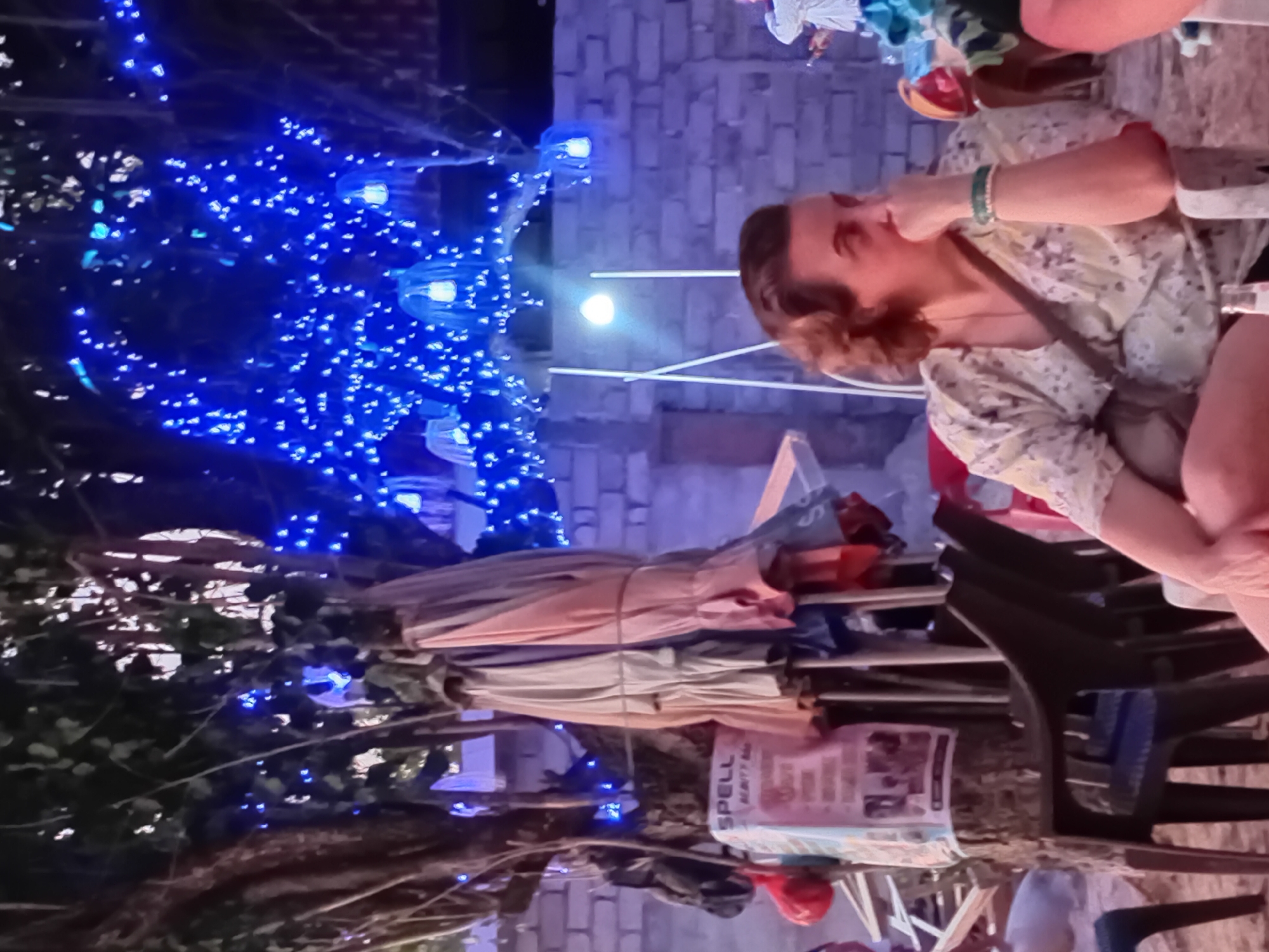 In Kuta, Indonesia, at night, a woman with short, wavy brown hair is seated in a chair, looking to her right with her right hand touching her chin. She is wearing a light-colored floral-patterned shirt and a green beaded bracelet on her left wrist. The background features a brick wall adorned with blue string lights, some of which are shaped like jellyfish. Several closed umbrellas are leaning against a tree to the left, near a sign that reads "SPELL BEAUTY BAR." Other people and chairs are visible in the periphery.