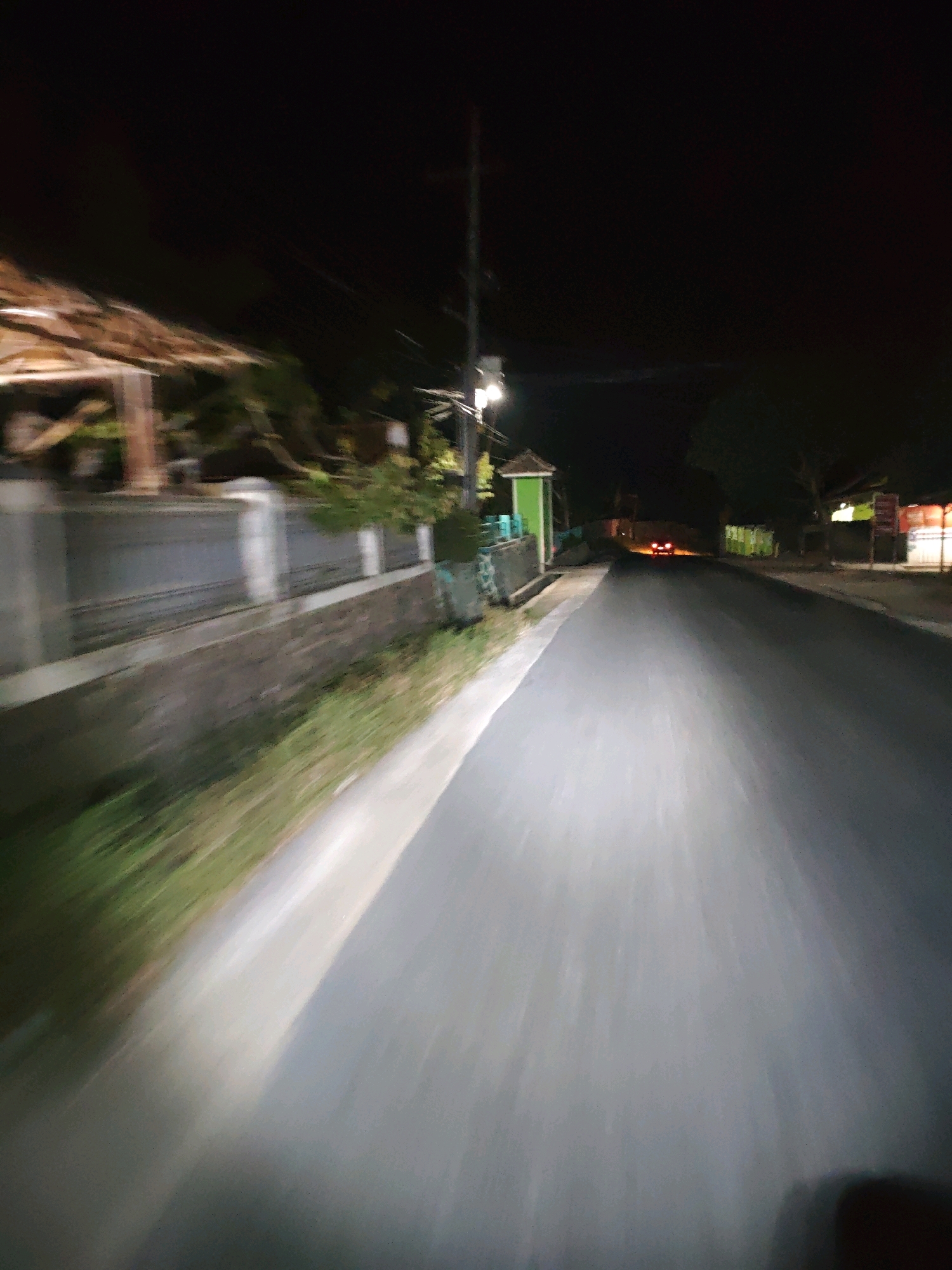 The image depicts a nighttime view of a street in Sukarame, Indonesia, likely from a moving vehicle. The road surface is dark and appears to be asphalt. A bright beam of light, presumably from headlights, illuminates the foreground, casting a strong glow on the road ahead. To the left of the road, there is a low wall and a grassy area with vegetation. Beyond the wall, glimpses of buildings with thatched roofs and structures with green accents are visible, suggesting a residential or commercial area. In the distance, the red taillights of a vehicle can be seen on the road. Streetlights are present, one of which is clearly visible on the left side of the road, providing illumination. The overall impression is that of a quiet street scene at night.