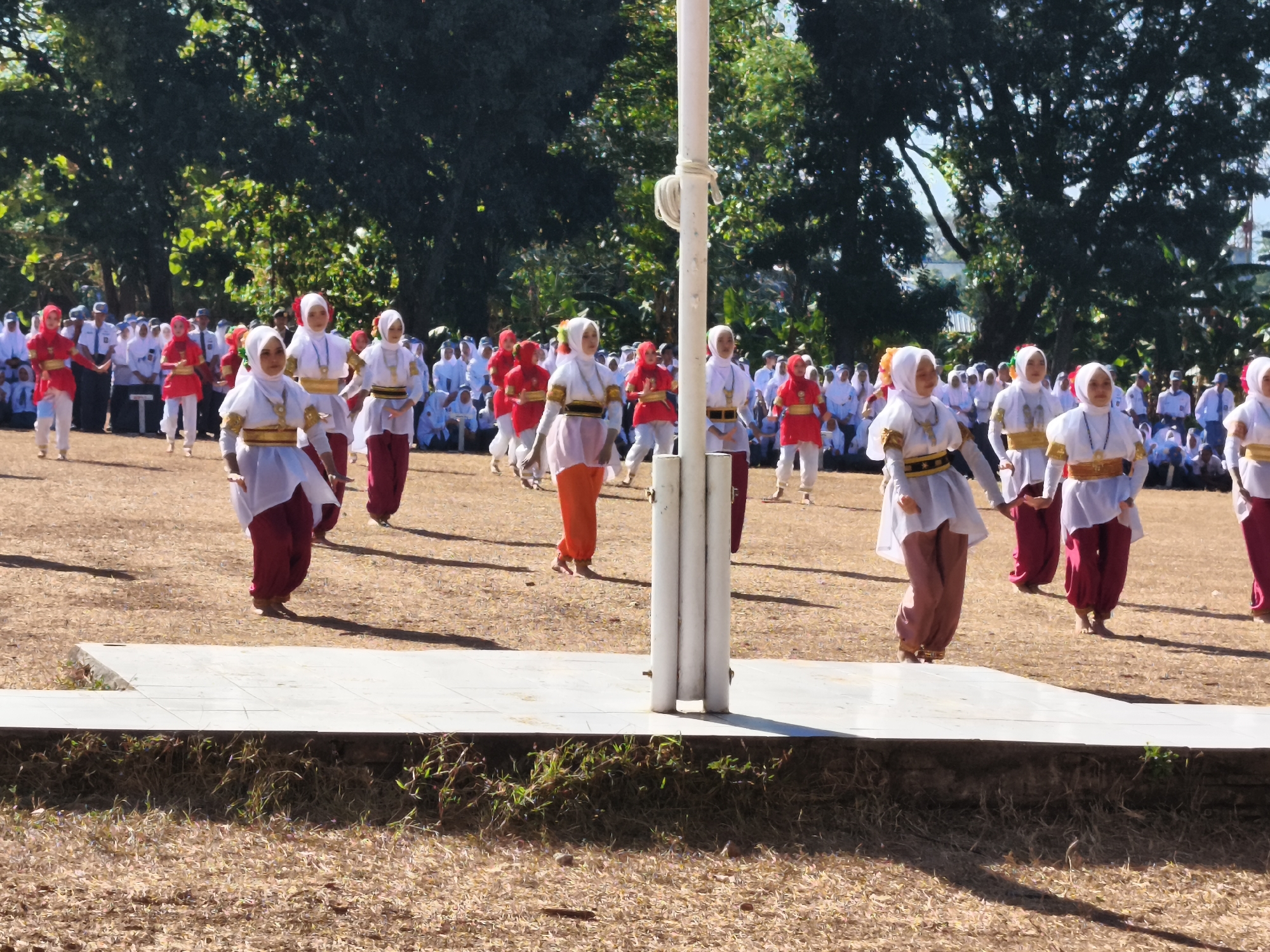 The image captures an outdoor performance by young women in traditional attire, likely during a cultural event or ceremony. The performers are dressed in brightly colored garments, with many wearing white tops and red or orange pants, and headscarves. Some have gold or black sashes around their waists. They are in motion, suggesting a dance or choreographed routine. In the background, a crowd of spectators, many of whom are also dressed in white, are seated and observing the performance. The setting appears to be a grassy field with trees lining the perimeter, indicating a park or an open public space. The presence of a flagpole suggests a formal event, possibly related to a flag-raising ceremony. Without explicit visual cues pointing to the city of Makassar, Indonesia, it is not possible to definitively place the location. However, the attire and the presence of many individuals in white suggest a cultural or religious event common in Indonesia.