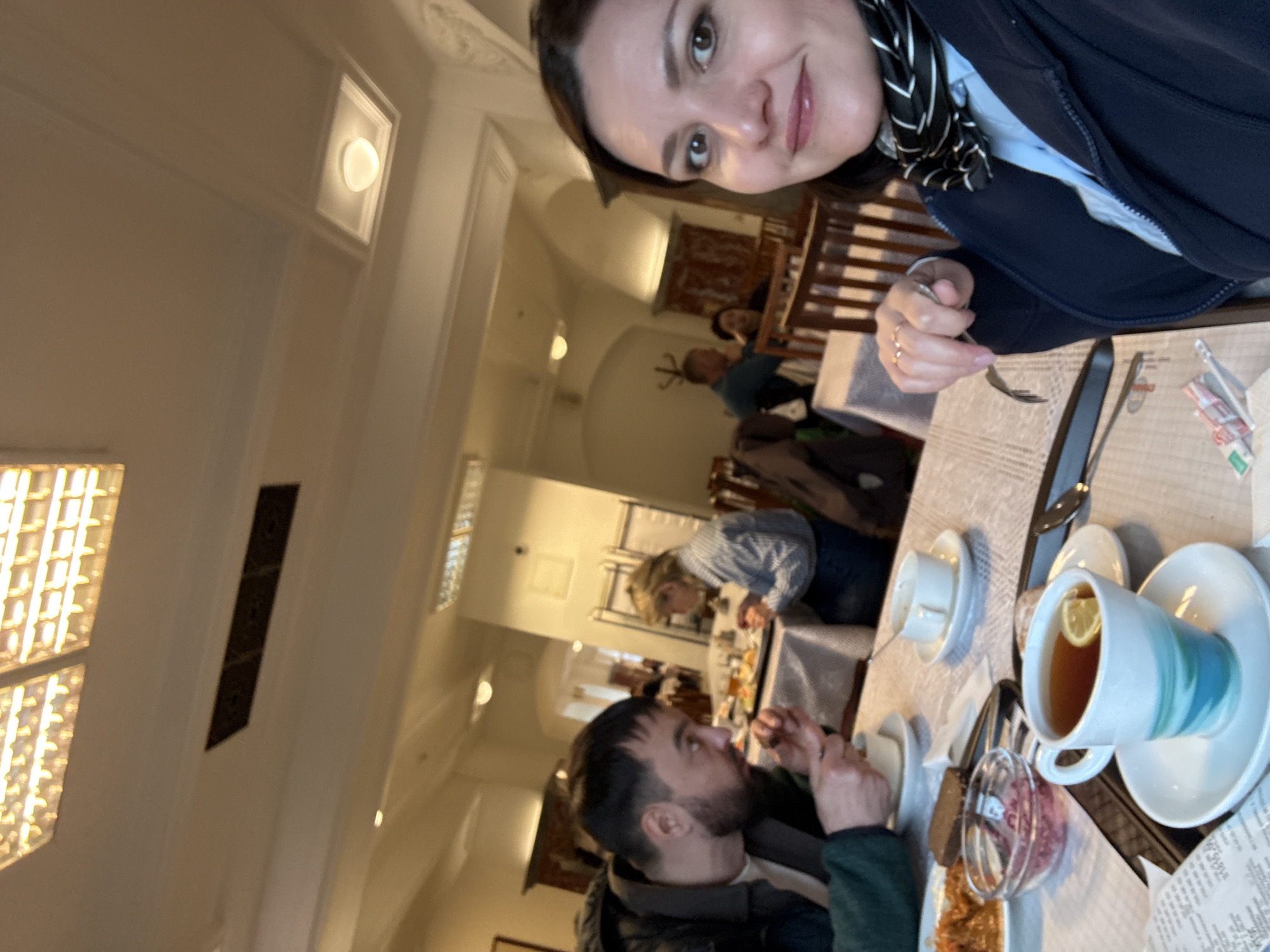 The image is an eye-level photograph showing a woman and a man at a table in a dining room. The woman, who seems to be holding the camera, has brown hair, light skin, and is wearing a dark jacket and a black and white scarf. She is smiling and looking at the camera. She is holding a small fork in her right hand.
The man at the table has dark hair and a beard. He is wearing a dark jacket. The two people are seated at a table set with teacups, saucers, a spoon, and a serving dish with some type of food.
In the background, other people are seated at tables in the dining room. The room has a high ceiling, white walls, decorative lighting, and arched doorways.
Based on the interior design and context clues, it is possible that the location could be in the city of Moscow, Russia.