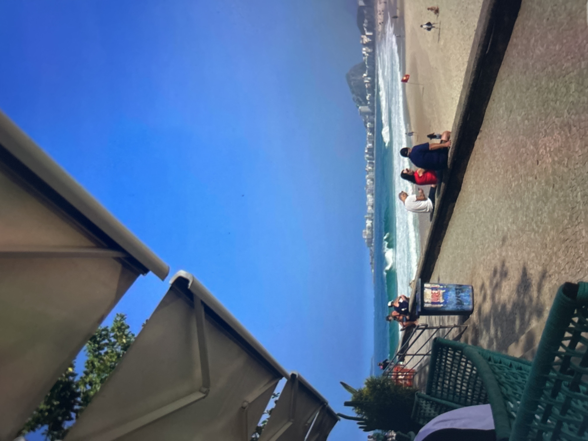 The image shows a sunny day at a beach in an urban setting. The foreground features a green metal bench and a concrete barrier, with a blue trash can in the middle. A white awning extends from the top left, providing shade.
Beyond the barrier, a sandy beach stretches out, populated by several people. Some are sitting on the barrier, facing the ocean. Others are walking or relaxing on the sand. The ocean is a vibrant turquoise, with waves rolling onto the shore.
In the background, a city skyline is visible, with numerous buildings and a prominent mountain. The sky is a clear, bright blue. The overall scene suggests a leisurely day at a popular beach destination.