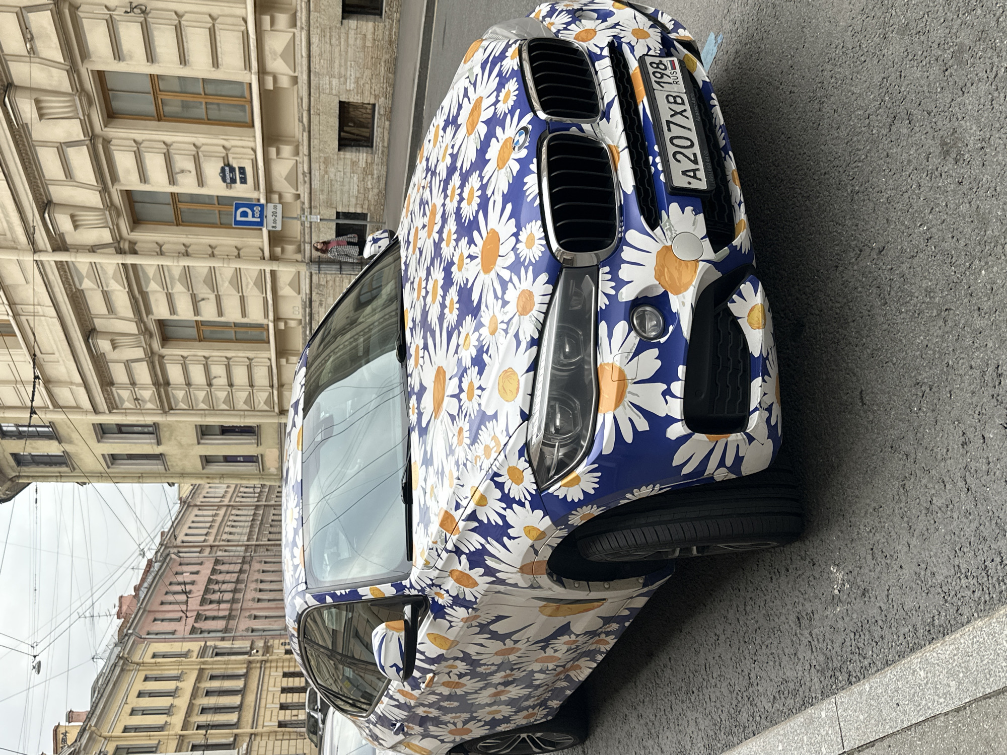 The image showcases the front of a blue BMW car adorned with a striking daisy floral print, parked on a city street. The car's license plate reads "A207XB 198 RUS," indicating it is registered in Russia. The background features a cityscape with buildings exhibiting various architectural styles. A blue "P" parking sign is visible, and a person is walking in the vicinity of the car. The location is identifiable as Saint Petersburg, Russia.
