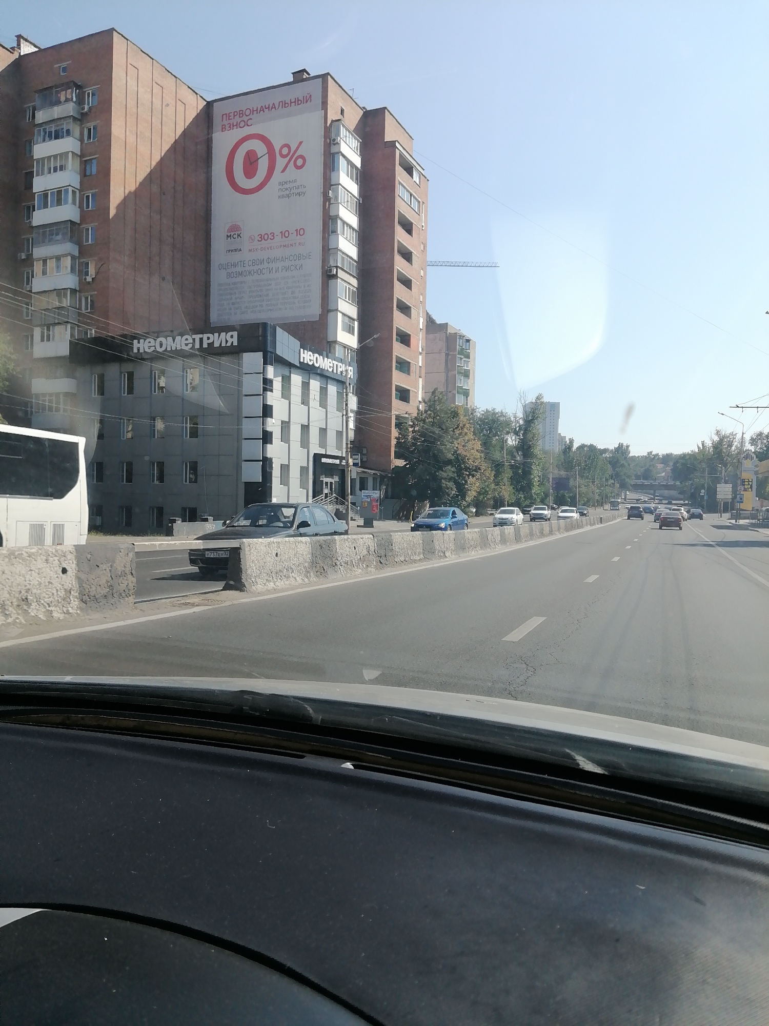 The image captures a view from inside a vehicle, looking out at a city street in Rostov-on-Don, Russia. In the foreground, the dashboard of the vehicle is visible. The street itself is paved, with multiple lanes of traffic. Several cars are present on the road, including a dark-colored sedan in the middle lane and a blue sedan further down. Concrete barriers line the side of the road.
On the left side of the street, a multi-story building with a brick facade is prominent. A large advertisement is displayed on this building, featuring a red circle with "0%" inside and text in Russian, including "ПЕРВОНАЧАЛЬНЫЙ ВЗНОС" which translates to "initial payment" or "down payment." Below this, the company name "МСК" and a phone number are visible. Further down, a modern-looking building with "НЕОМЕТРИЯ" written in large letters on its facade is situated. A white bus is also parked on the left.
The background shows more buildings, trees, and a clear blue sky. The overall impression is of a typical urban street scene on a sunny day.