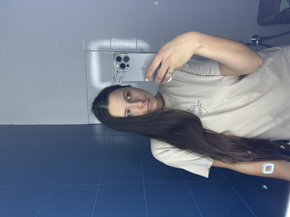 A female subject is captured in a mirror selfie within an indoor setting. She possesses long, dark hair, parted centrally, and wears a beige short-sleeved t-shirt featuring a graphic design on the chest. A square, white-outlined patch is visible on her left upper arm. Her right hand, adorned with a multi-banded silver ring on the ring finger and white-painted fingernails, holds a light-colored smartphone encased in clear plastic with a stylized animal head design around its triple camera module. The subject's expression is neutral, with her gaze directed to her left (viewer's left). The background consists of tiled walls: dark blue rectangular tiles are on the left side of the reflection, and light gray rectangular tiles are on the right, meeting at a vertical seam. A light source reflects off the lighter tiles. A portion of a dark, woven basket or container is visible in the lower right. The precise location within Shul'gino, Russia, is not ascertainable from the visual evidence.