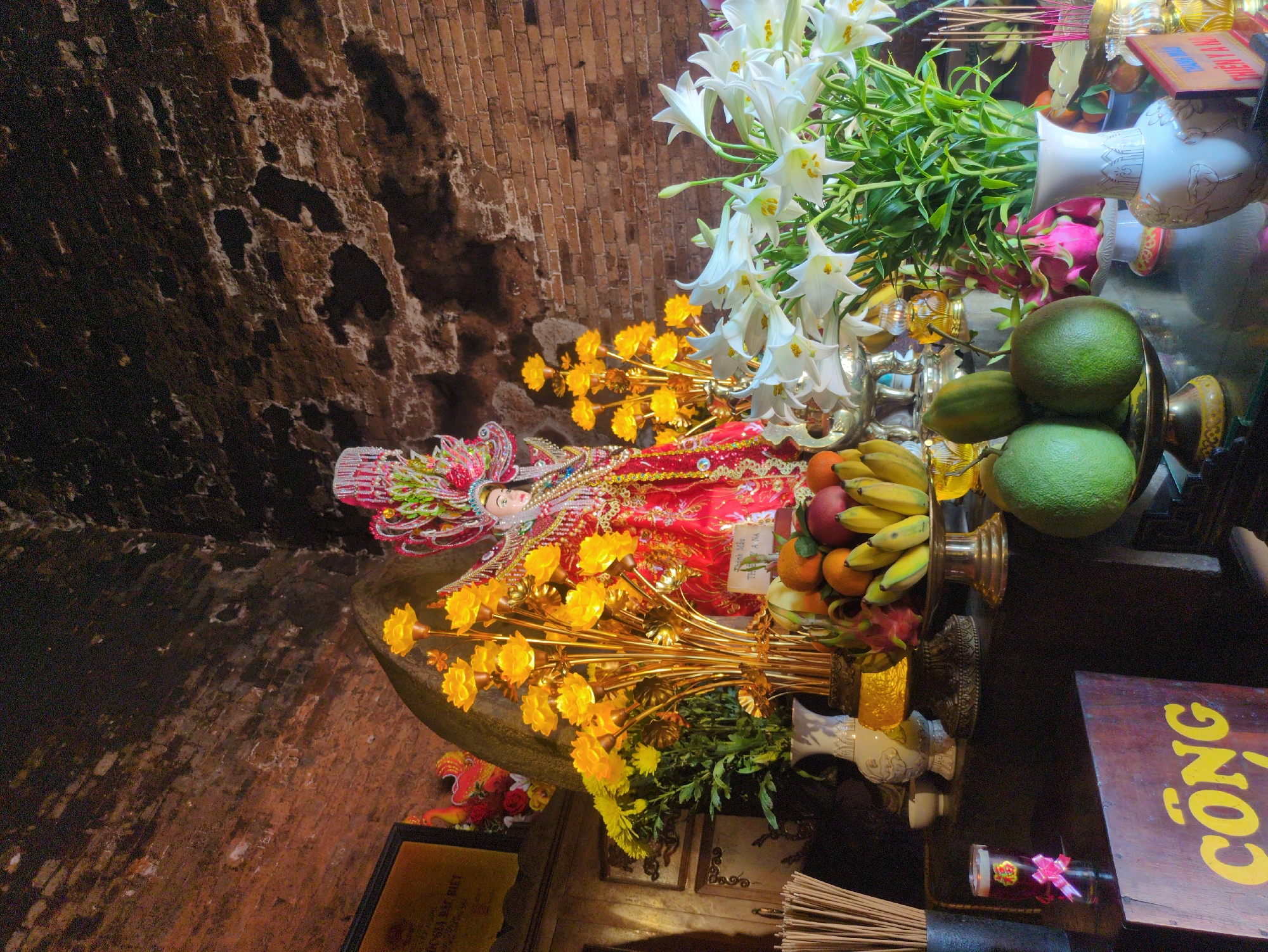 The image shows a shrine inside a structure with a brick wall. The shrine includes a statue dressed in red and adorned with yellow flowers, white lilies, and various fruits like bananas, oranges, and pomelos. There are also vases and a wooden sign with the word "CÔNG" written in yellow. The location is likely Tuy Hòa, Vietnam, given the Vietnamese text on the sign.
