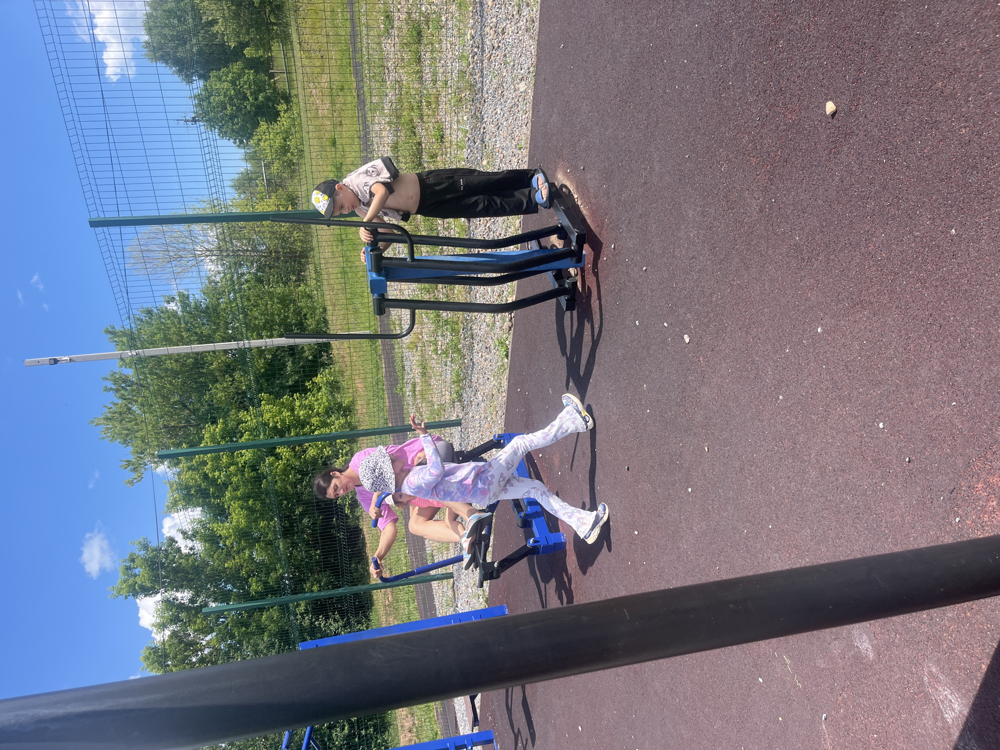 This image captures two individuals, likely children, engaging in physical activity on an outdoor gym apparatus. The scene is set in a park or recreational area with a dark red, rubberized ground surface. A green metal fence, partially obscured by vegetation, borders the area, with trees and a bright blue sky with scattered clouds in the background.
The primary focus is on two figures on a blue and black exercise machine. One person, appearing to be a young boy wearing a black baseball cap and black pants, is in a plank-like position, holding onto the machine's handles. He is shirtless, revealing his stomach. The second individual, a younger child dressed in a pink t-shirt and white patterned leggings, is seated on a different part of the apparatus, also holding onto handles. Their posture suggests they are actively using the equipment.
The lighting indicates it is daytime, likely sunny, given the clear sky and shadows cast by the gym equipment and the people. The overall mood appears to be one of active recreation and perhaps a shared moment of exercise between the two individuals. The presence of a fence, trees, and an open outdoor space suggests a public park or sports ground in a rural or suburban setting. There is no visible text in the image.