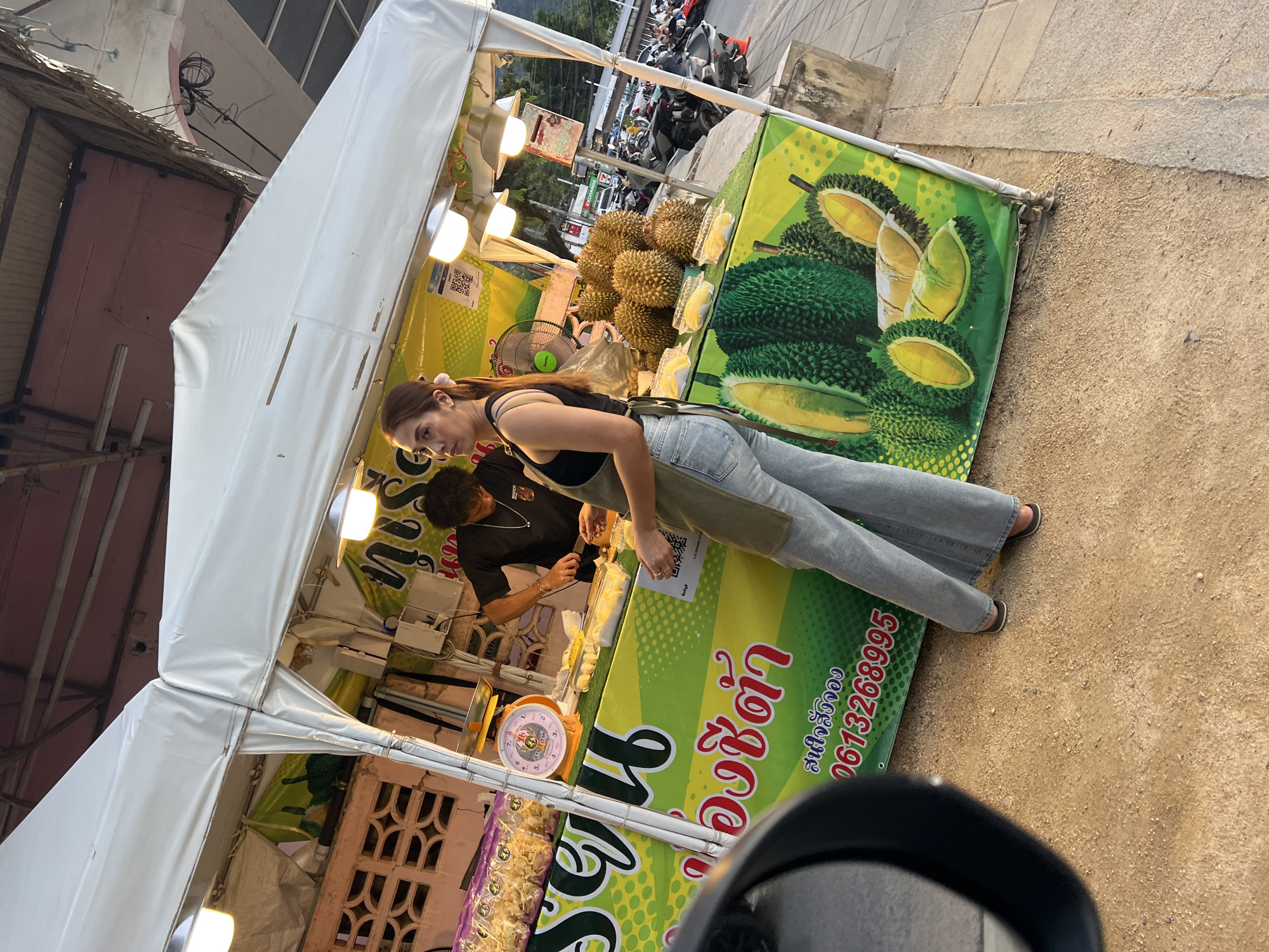 A street scene in บ้านบางรักษ์, Thailand, depicts a durian vendor operating from a roadside stall beneath a white tent. A young woman with long brown hair, wearing a black tank top, light blue jeans, and an apron, stands at the counter, appearing to engage with a customer, a man wearing a black shirt. Numerous durians are stacked near the front of the stand. A green and yellow sign displaying text in Thai and images of durian adorns the stall's front. A scale sits on the counter amidst the durians. In the background, various motorcycles are parked, and part of a vehicle mirror is visible in the foreground.