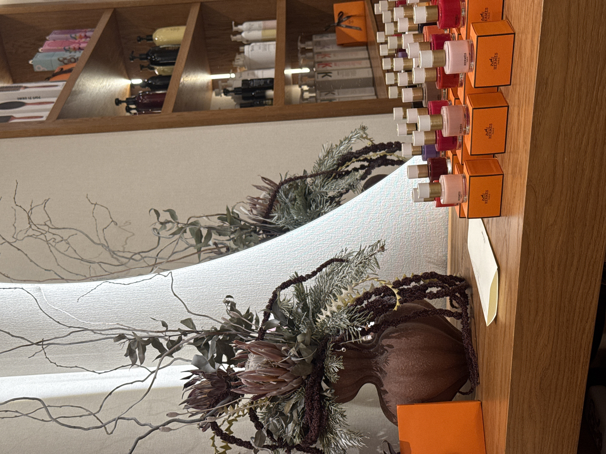 This image captures an interior scene, specifically within what appears to be an upscale beauty salon or boutique in Yerevan, Armenia. The focal point is a wooden counter or display table, featuring a collection of luxury beauty products and decorative elements.
On the left side of the counter, a large, dark reddish-brown, organic-shaped vase holds an elaborate dried floral arrangement. This arrangement consists of large, muted pink-brown protea-like flowers, dried eucalyptus leaves, coniferous branches, and cascading dark reddish-brown amaranth-like foliage. To the immediate left of the vase, an orange square box, likely from Hermès, is positioned. A small, light-colored, folded paper or envelope lies flat on the counter in front of the vase.
Across the right side of the counter, numerous small glass bottles of nail polish are meticulously arranged in rows. Each bottle features a white cap with a gold band and is displayed on its individual orange square box, clearly branded "HERMÈS PARIS." The visible polish colors include various shades of pink, red, purple, and neutral tones.
In the background, on the left, a textured light-colored wall or fabric screen is visible, partially adorned with thin, bare, twisting branches as decorative elements. On the right, a tall, light-brown wooden shelving unit extends vertically. The shelves are stocked with various beauty and hair care products. The upper shelves display retail boxes, some featuring text such as "FOR HER HAIR." The middle and lower shelves contain an assortment of bottles, primarily professional-sized, in dark colors (red, black, yellow) and lighter shades, many with pump dispensers. Some bottles on the lower shelf clearly show "KK" branding, indicative of Kérastase products. An additional orange Hermès gift box, tied with a dark ribbon, is visible on one of the lower shelves. No individuals are present in the scene.