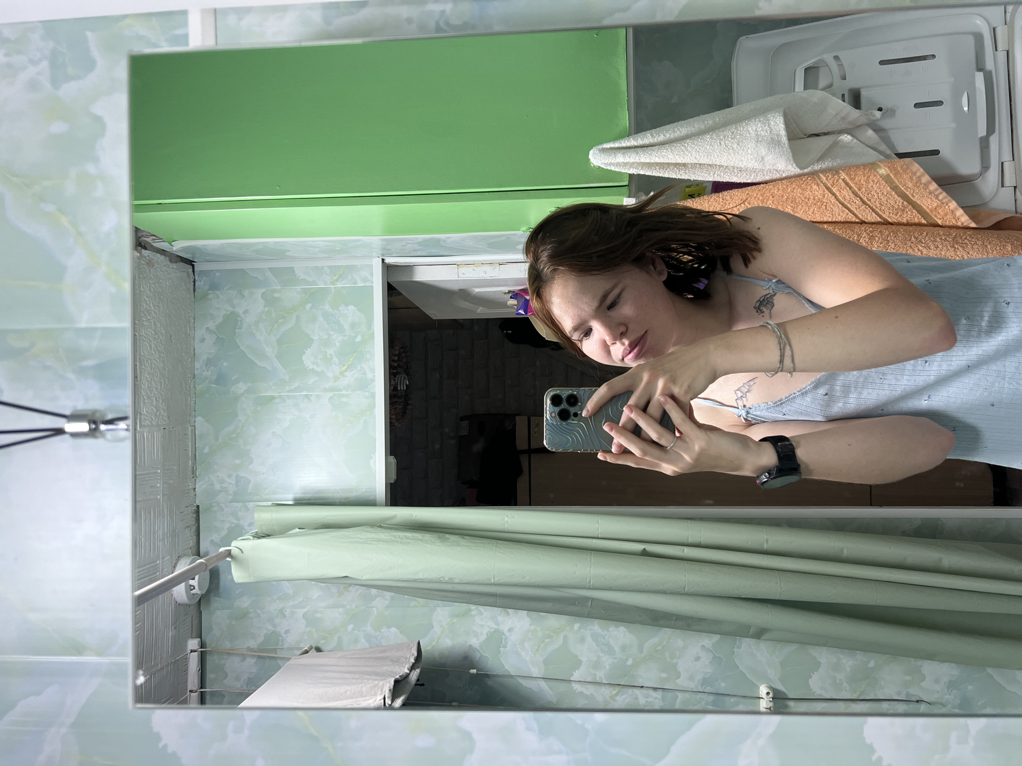 The image shows a young woman taking a selfie in a bathroom. She is wearing a light blue tank top and has brown hair. She is holding a smartphone in her right hand. The bathroom has light green marbled walls, a light green shower curtain, and a green cabinet. There is a white towel and an orange towel hanging on the right side of the image. A white washing machine is visible on the far right. The scene appears to be inside a residential apartment in the city of Chaikovsky, Russia.