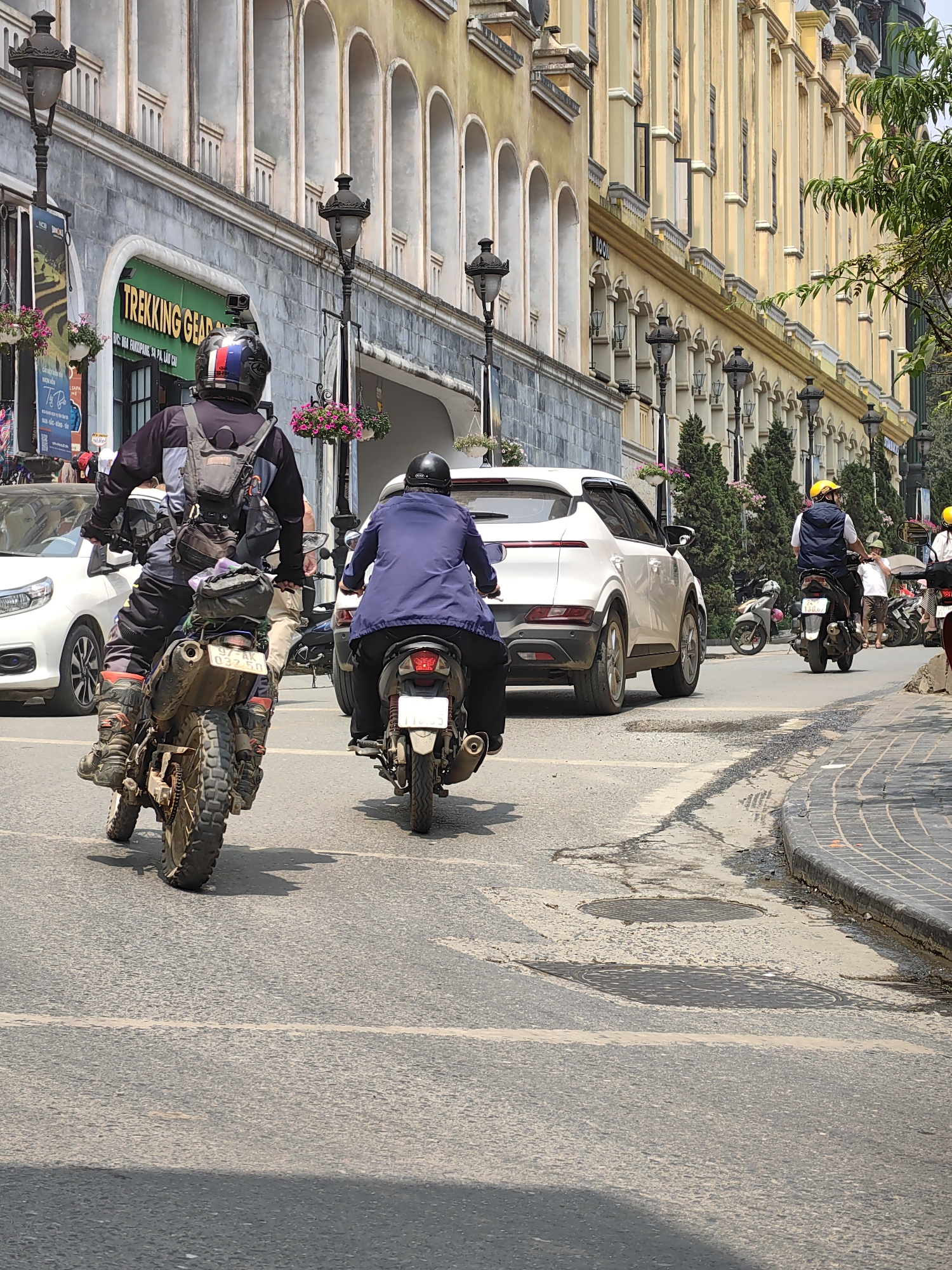 The image depicts a street scene in Sa Pa, Vietnam, under bright daylight. In the immediate foreground, two individuals are riding motorcycles away from the viewer. On the left, an individual operates a dirt-style motorcycle, heavily laden with gear bags attached to the rear. The rider wears a black and gray long-sleeved jacket, dark pants, and dirt bike boots, topped with a black helmet featuring red, white, and blue stripes. The motorcycle's rear license plate displays "97E-AK 037.50." To the right and slightly ahead, a second individual rides a dark-colored scooter or smaller motorcycle, wearing a blue jacket and a black helmet. Its visible rear license plate is "116.35."
The street is paved with asphalt, showing signs of wear, patches, and two circular manhole covers. A white lane line is partially visible in the mid-ground. To the far right, a tiled curb separates the road from a sidewalk.
On the left side of the street, a multi-story building features a gray, stone-textured lower facade. A prominent green sign for "TREKKING GEAR" is visible. Above this, the building transitions to a lighter color with a series of arched windows. Multiple ornate black streetlights, each with a hanging basket of pink flowers, are affixed to the building.
Further down the street, a white SUV is positioned in traffic, partially obscuring another scooter rider wearing a blue vest and a yellow helmet. A white Honda CRV is also partially visible on the left side, behind the lead motorcyclist. On the right, a long building with a distinctive yellow and cream facade extends into the distance, characterized by numerous arched windows and vertical architectural details. Parked motorcycles and pedestrians are visible along the right side of the street, including one individual in a light-colored shirt and hat. Green foliage from trees is visible on the far right. Shadows are cast across the street, consistent with sunny conditions.
