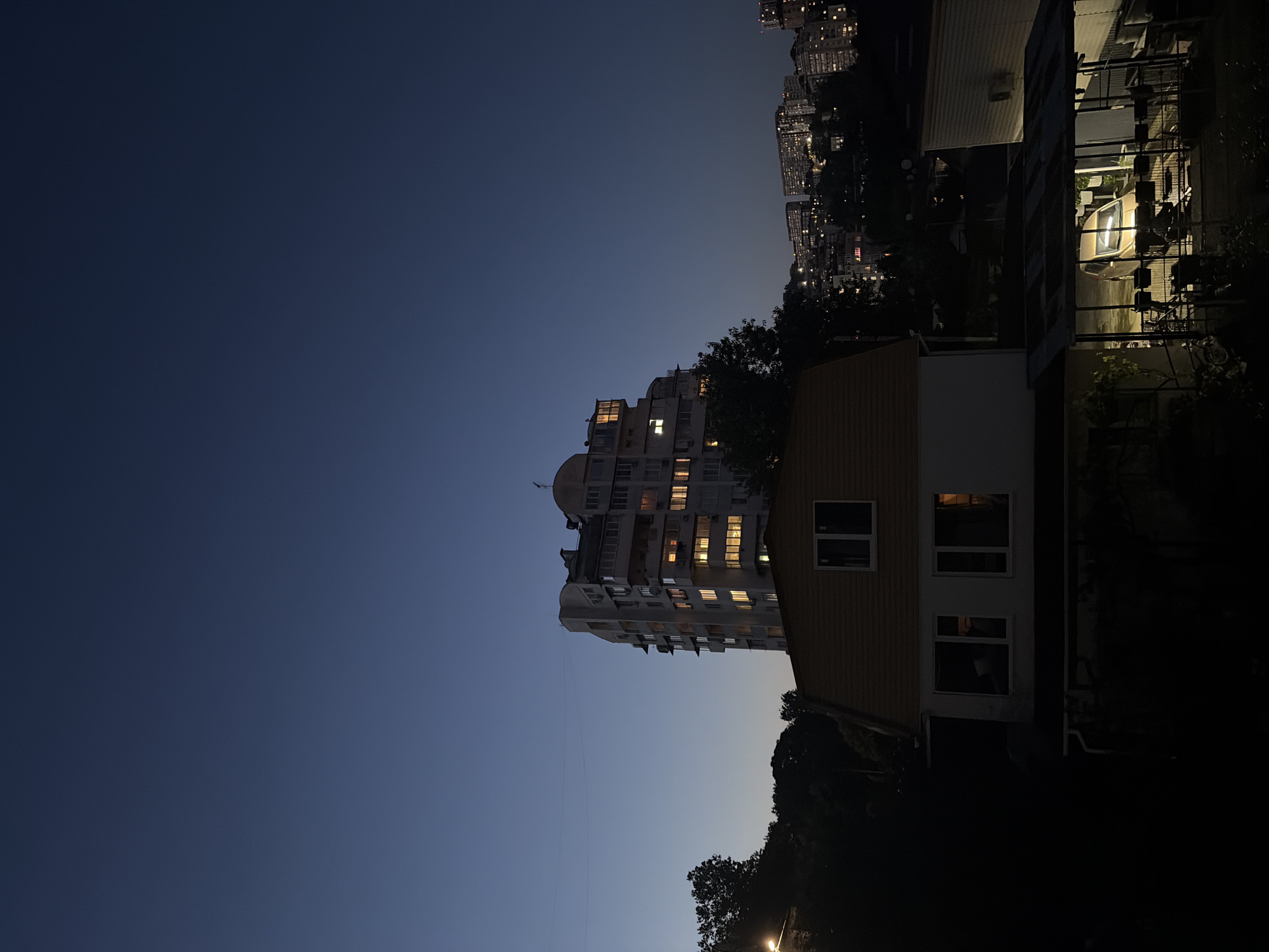 The image captures a nighttime or late dusk scene in Sochi, Russia, depicting an urban landscape with residential buildings and natural elements under a deep blue sky. In the mid-ground, a multi-story, light-colored apartment building dominates the center, featuring numerous illuminated windows across its various floors, indicating active occupancy. The building exhibits a somewhat complex architectural structure with balconies, external air conditioning units, and an antenna on its roof.
In the immediate foreground, a two-story house with a distinctive gabled (mansard) roof is positioned, its upper facade clad in orange-yellow siding and its lower half in white. Several windows are visible on the house, with some reflecting ambient light. To the far right, a covered structure, possibly a carport or patio, shelters a light-colored sedan (gold or beige) under artificial illumination. Below this structure, parts of a dark metal railing or fence are visible, along with what appears to be a bicycle.
The left and right sides of the foreground and mid-ground are characterized by dark, dense tree foliage, suggesting a verdant environment. On the far left, a bright, circular light source is partially visible amidst the trees. In the distant background, a sprawling cityscape extends across a hillside, marked by a multitude of small, scattered lights from buildings and possibly streetlights, indicative of a densely populated urban area built on varied topography. A thin power line is discernible stretching across the sky in the upper-left quadrant. No individuals or specific actions are identifiable in the scene.