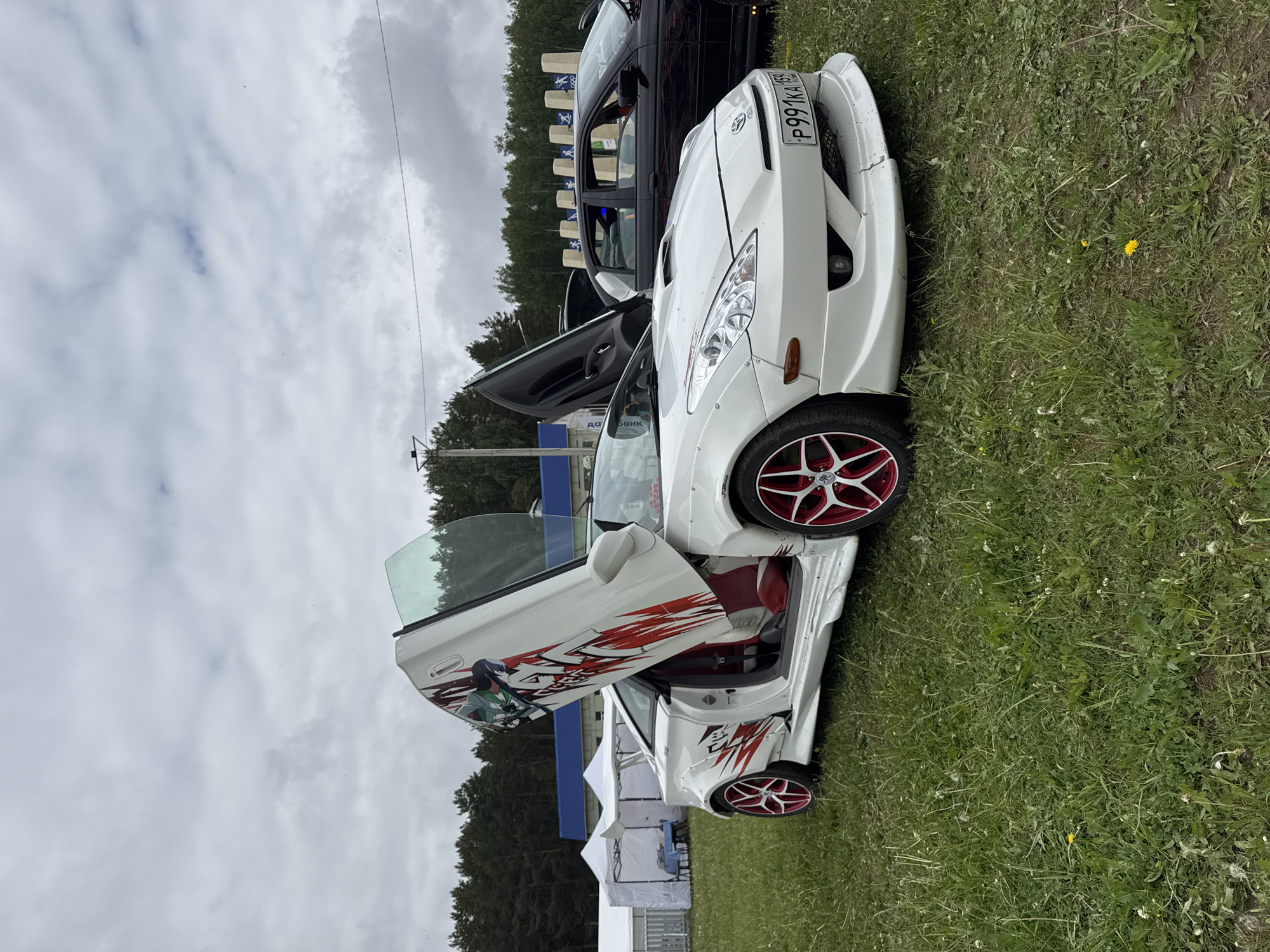 The image depicts two customized cars parked on a grassy field, presumably in an outdoor area in Chaikovsky, Russia. The primary focus is a white car with aftermarket modifications, including scissor doors featuring anime-style graphics in red and black. The car also has red and silver rims, and a prominent front bumper modification. The license plate reads "P991KA 163". The second car is black, parked adjacent to the white one, with its door also open. Tents are visible in the background, indicating the setting might be a car show or event. The sky is overcast.