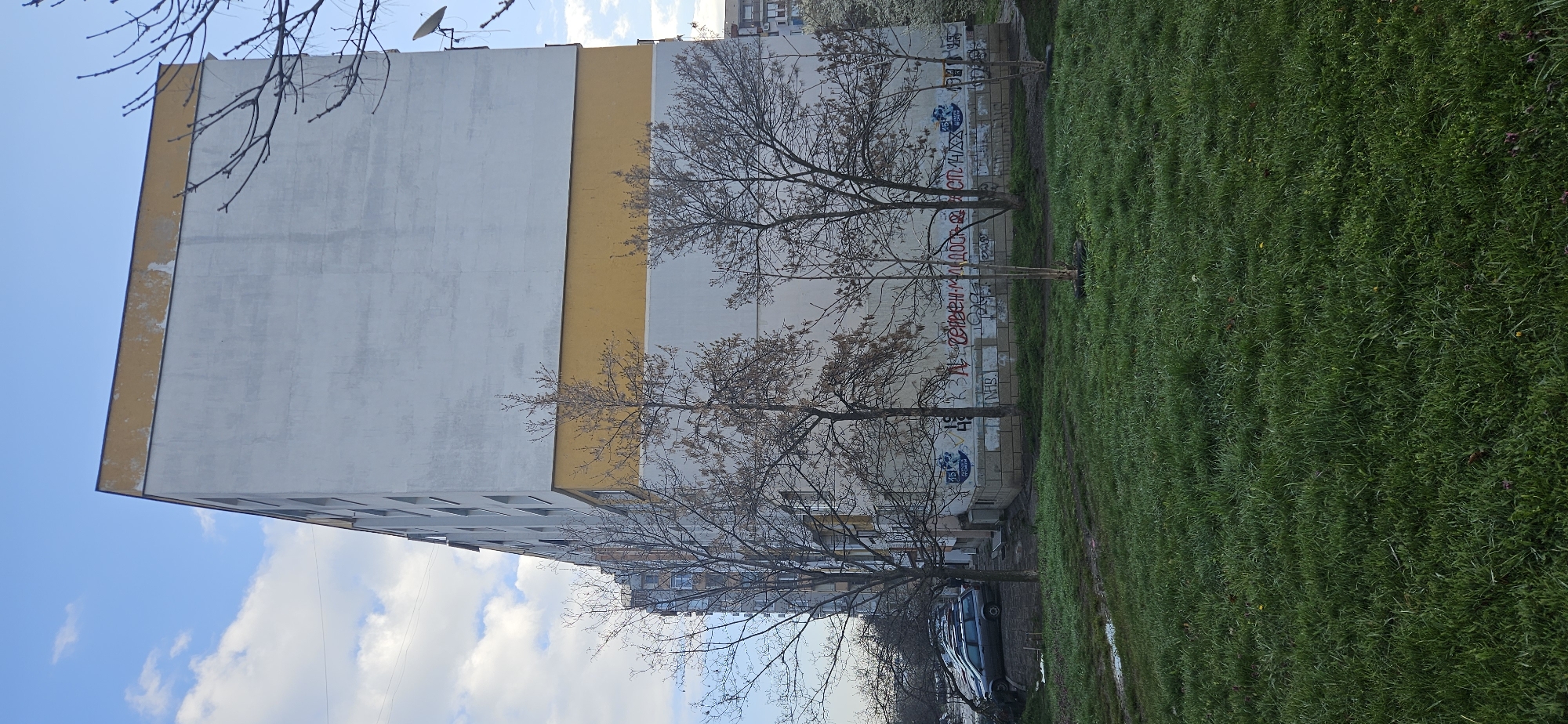 The image depicts a multi-story building in Sofia, Bulgaria. The building has a light gray and yellow facade, with graffiti visible on the lower portion. Several bare trees stand in front of the building. A patch of green grass is adjacent to the building and stretches into the foreground. A car is parked near the building. The sky is partially cloudy with a visible blue sky. A satellite dish is mounted on the building's roof.