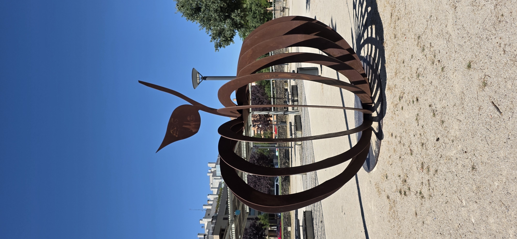 The image shows a large, abstract sculpture of an apple made of rusted metal. The sculpture is located in a public outdoor space with a sandy ground. In the background, there are buildings with white facades, a lamppost, trees, and covered seating areas. The sky is clear and blue. The sculpture is positioned in the foreground, dominating the frame. The overall scene suggests a park or a public plaza in a city. This sculpture is located in the Parque Miguel Servet in Huesca, Spain.