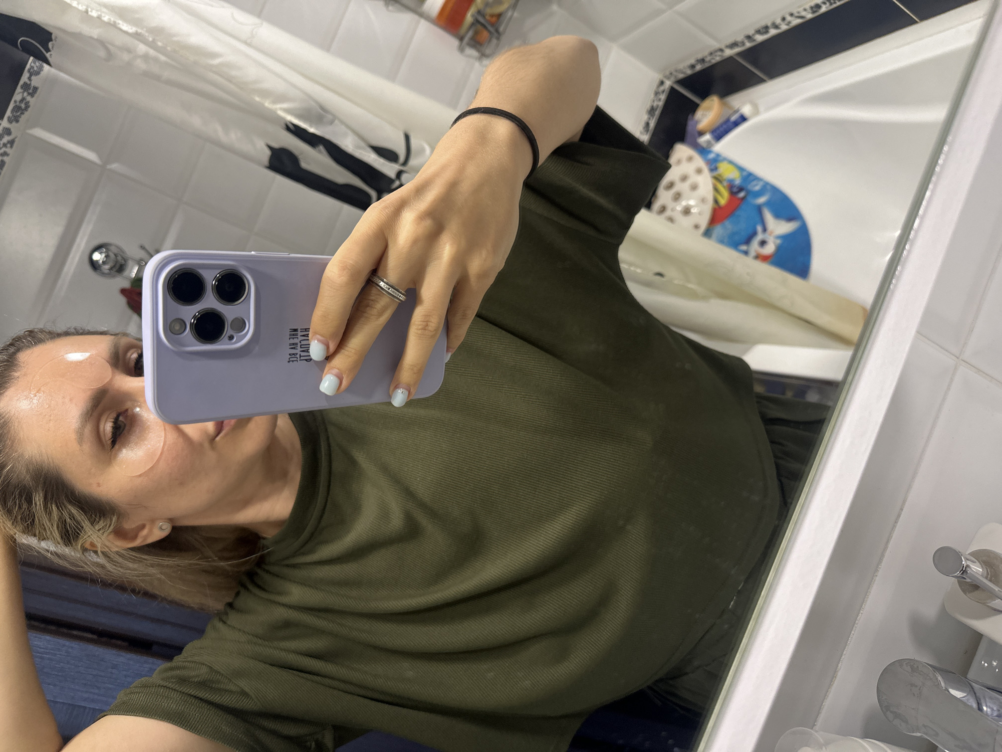 A woman with blonde hair pulled back is taking a selfie in a bathroom mirror. She is wearing a green t-shirt and has eye patches on. She is holding a light purple phone in her right hand. The bathroom has white tiled walls and a bathtub with a child's bath mat visible. A faucet and soap dispenser are on the counter in the foreground. The scene appears to be within a residential setting in Novoaltaysk, Russia.