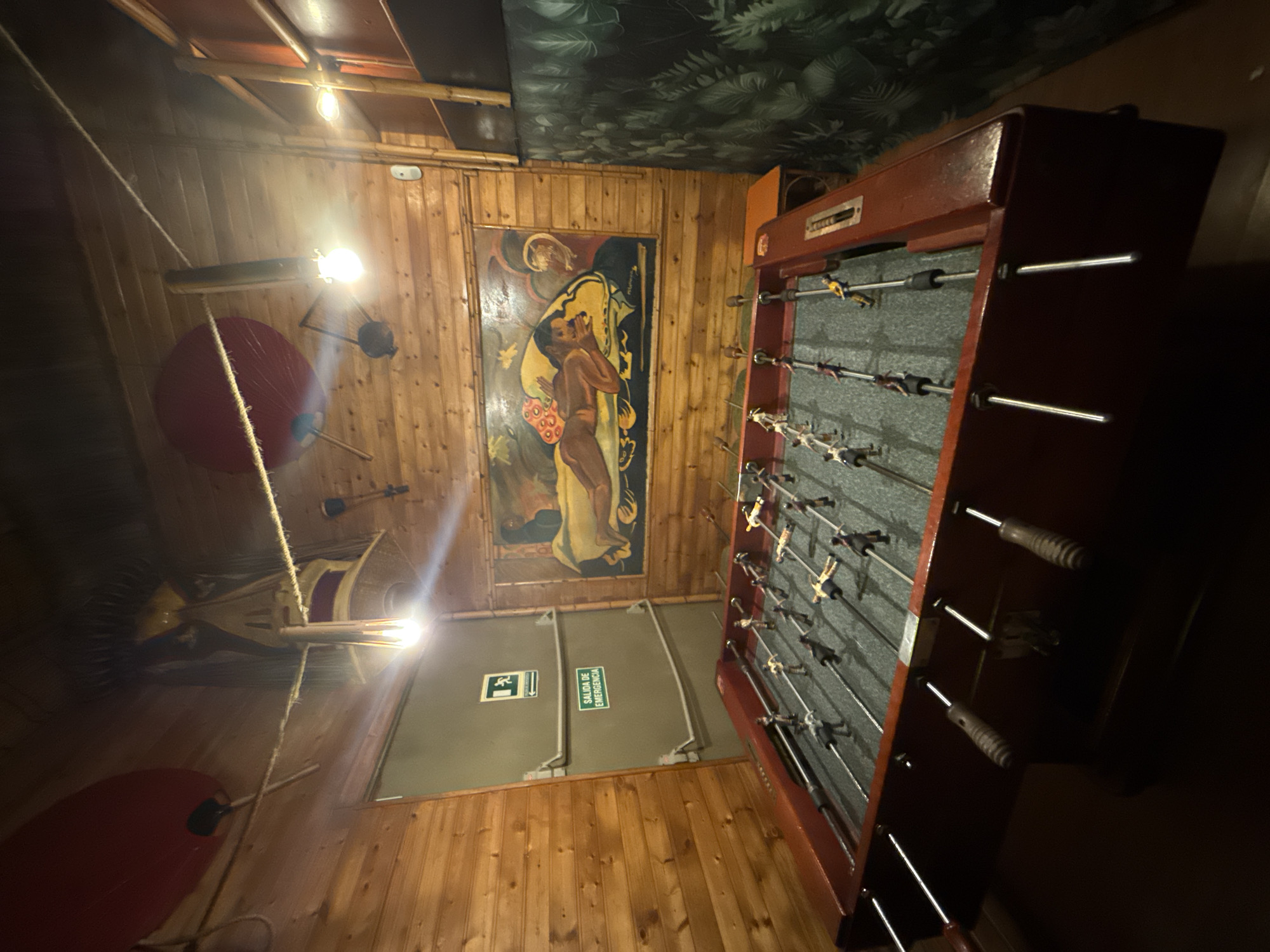 The image shows the interior of a building. The walls are wooden, and there is a painting on one wall. An emergency exit door is also visible. A foosball table dominates the scene. The ceiling is decorated with plant-themed wallpaper. The building is located in the city of Barcelona, Spain.