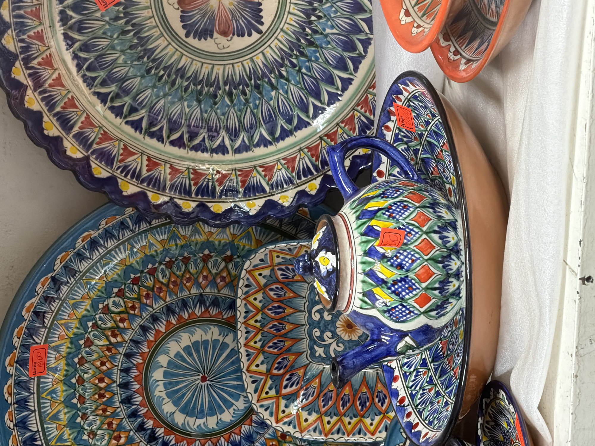 This image shows a display of intricately decorated ceramic tableware, likely from Central Asia, in what appears to be a market or shop. The central focus is a blue and white teapot with a colorful geometric pattern, placed on a matching decorative plate. Behind the teapot, several large, ornate ceramic plates are visible, featuring elaborate blue, white, and red designs. The plates have a scalloped edge and a concentric pattern. There are also smaller orange ceramic dishes stacked in the foreground on the right. Two red price tags are visible on the items; one on the large blue plate in the background reads "Цена 1800" and another on the plate beneath the teapot reads "Цена 1200". The items are displayed on a white, textured surface, possibly fabric, on a white shelf or counter. The lighting suggests an indoor setting. The image does not contain any people and there is no indication that this scene is located in Sochi, Russia.