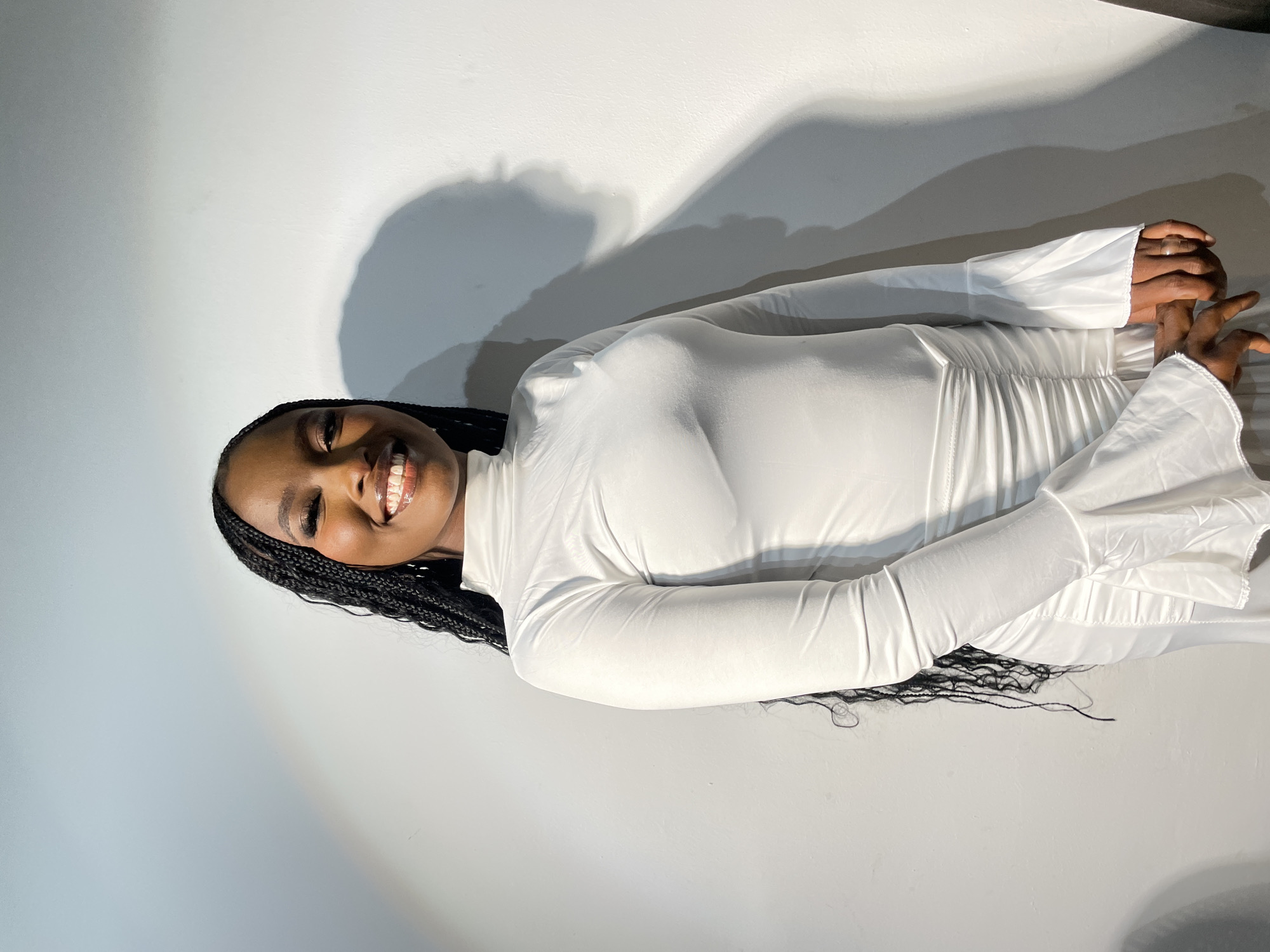 A young Black woman smiles at the camera while lying on her right side against a gray wall. She is wearing a white, form-fitting dress with long, flared sleeves. Her hair is styled in long braids. There is a shadow of a person or object cast on the wall behind her. There are no indicators to confirm this image was taken in Barakin Akawo, Nigeria.