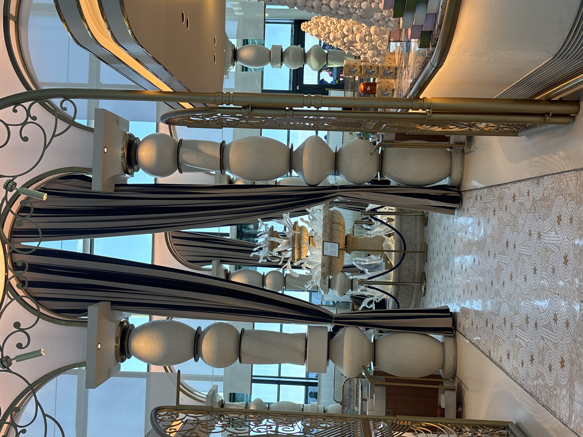 The image displays an interior scene, likely a hotel lobby or a high-end establishment, featuring ornate architectural details and decorative elements. The dominant colors are white, gold, and navy blue, creating a sense of opulence. Several large, white, urn-like structures with rounded, bulbous forms are positioned vertically, supporting golden railings and decorative scrollwork. These structures are interspersed with sections of navy and white striped fabric, possibly drapes or canopy elements.
In the background, a modern chandelier or art installation made of white, irregular shapes hangs from the ceiling, further enhancing the luxurious ambiance. There are glimpses of people in the background through glass partitions, suggesting it is an active public space. The floor is adorned with a patterned mosaic tile, adding another layer of intricate design. The lighting appears to be artificial, casting a warm glow on the surfaces. The overall impression is one of sophisticated interior design.
The scene appears to be set during the day, given the natural light filtering through what seem to be large windows or glass walls, though the primary illumination comes from interior lighting. No specific activity or event is clearly discernible, beyond the general presence of people and the implied use of the space as a reception or common area. The location context of Arnavutköy, Turkey, suggests a potentially waterfront or upscale urban setting. No visible text is present in the image.
