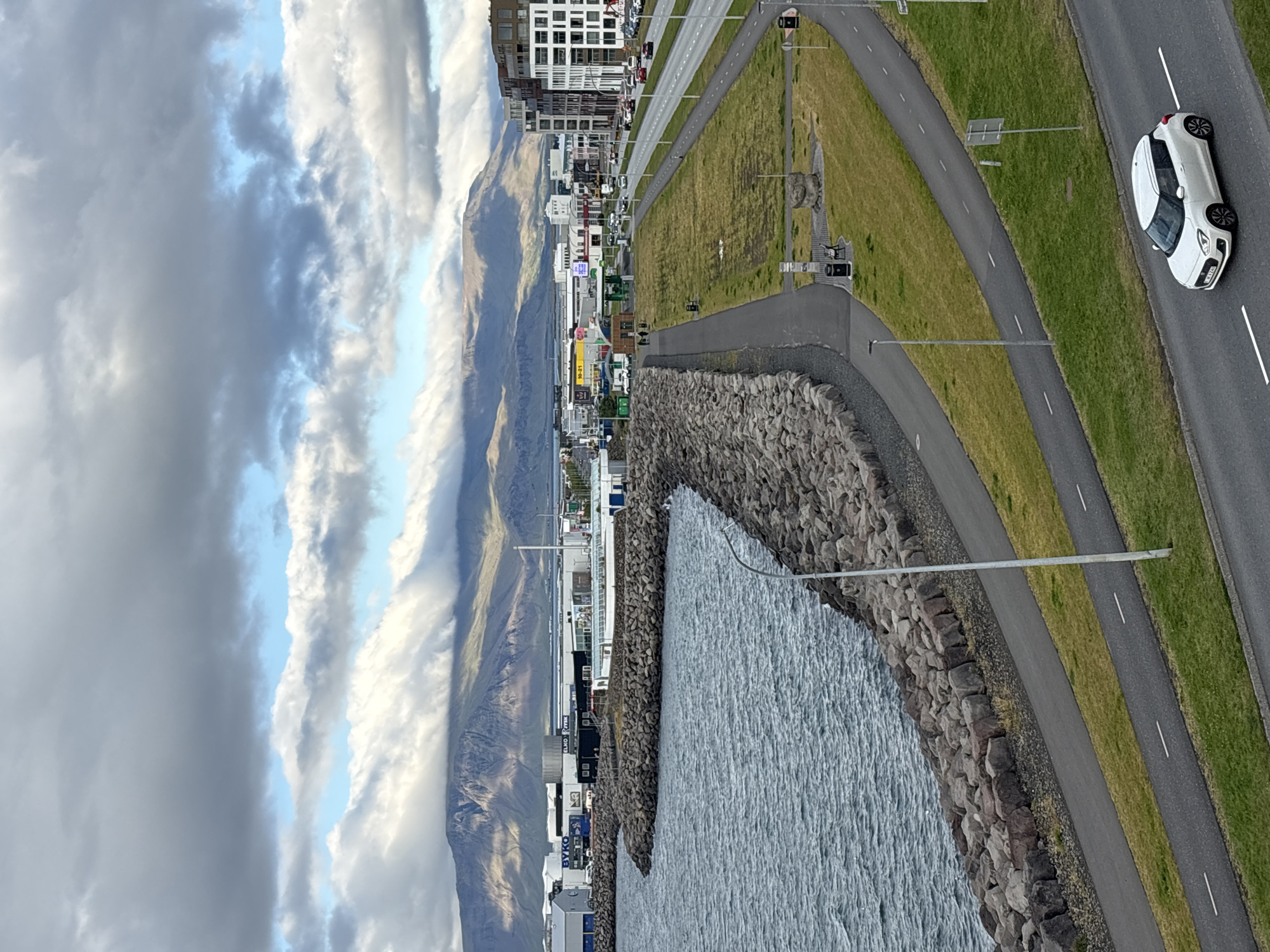 This is a medium-shot, eye-level, outdoor photograph taken in Reykjavik, Iceland, with a view overlooking a harbor, a paved pathway, and a road with a car. The sky is filled with dramatic clouds, with patches of blue sky visible. The foreground features a body of water on the left, bordered by a man-made rock wall. A paved walkway runs alongside the rock wall, and further inland, a grassy area is visible. A road is situated to the right of the grassy area, with a white car driving on it. In the background, there is a cityscape with various buildings and a large mountain range beyond. The overall impression is that of an urban waterfront area on a partly cloudy day.