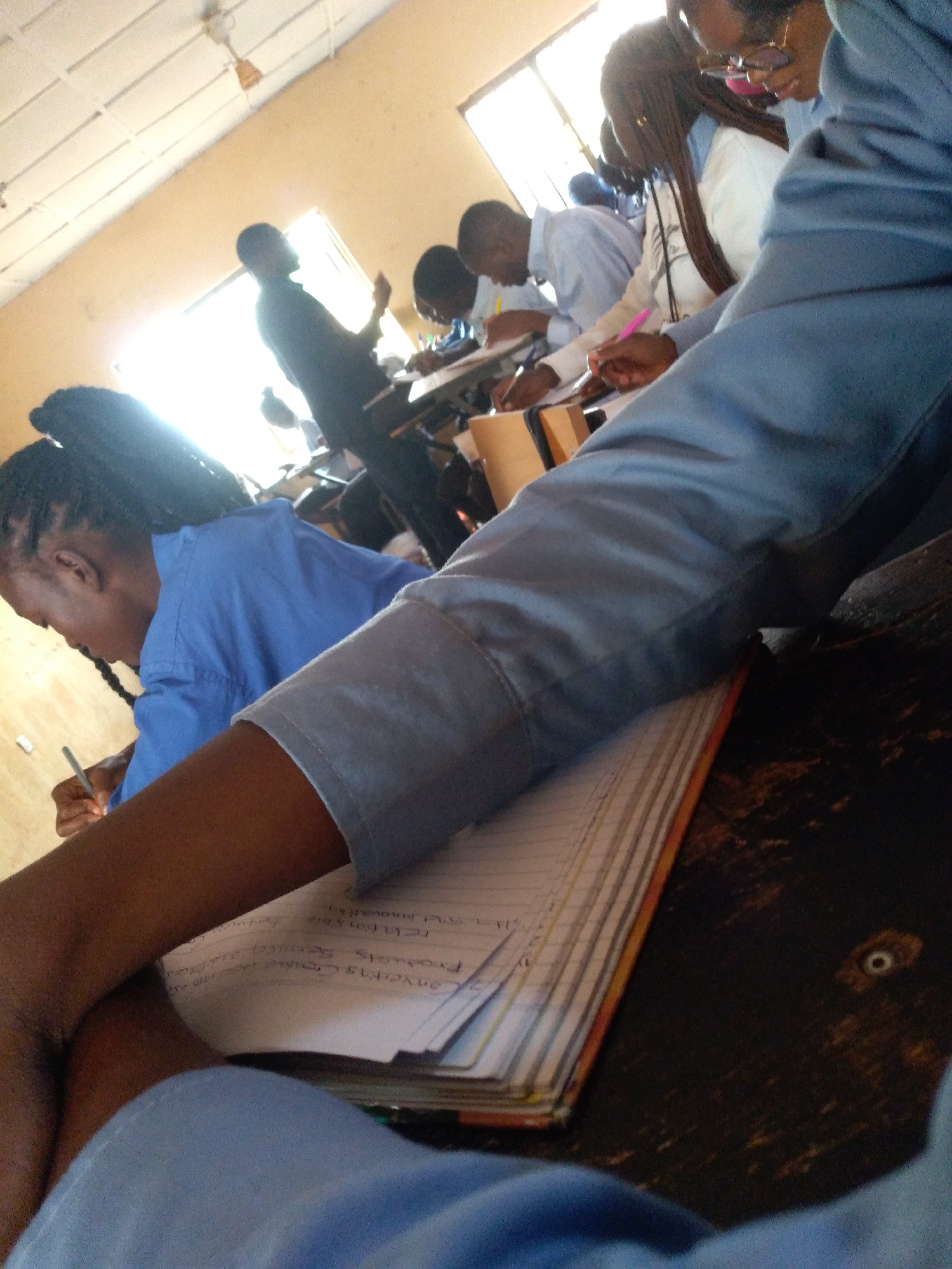 The image depicts a classroom setting in Jos, Nigeria, with several students seated at desks, engaged in writing or reviewing notes. The lighting suggests it's daytime, with natural light streaming in from a window. In the foreground, a student's arm and shoulder, clad in a light blue uniform, are prominent. This student is holding a pen and appears to be writing in a notebook that lies open on the desk. The notebook's pages are filled with handwritten text.
Further into the scene, other students are visible. One student, wearing a blue uniform, has their hair in braids and is also writing. Behind them, other students are seated, some leaning forward, suggesting concentration on their work. A male figure in a dark shirt stands near a window in the background, possibly a teacher or another student. The classroom environment is basic, with a tiled ceiling and yellow-painted walls. The desks appear to be made of wood. The overall impression is one of an academic environment in session.