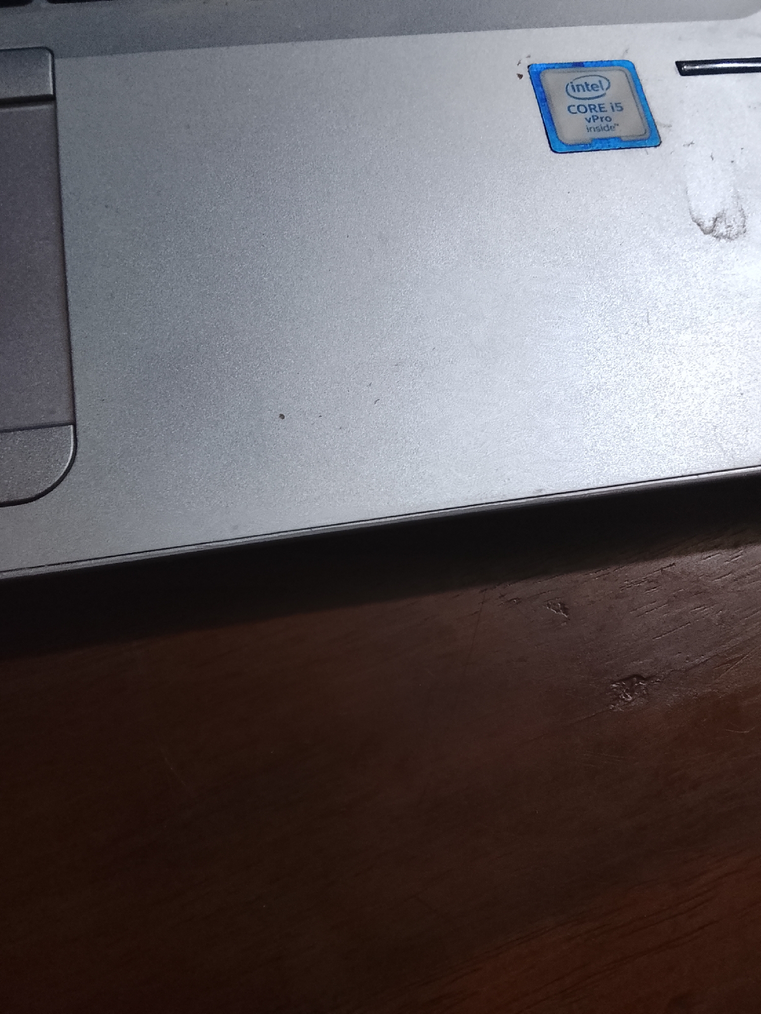 The image shows a portion of a laptop resting on a wooden surface. The laptop features a silver-colored casing with a trackpad on the left side and an Intel Core i5 vPro sticker on the upper right. The wooden surface is dark brown and appears to be a table. There are no people visible, and no specific actions are occurring. There is no information in the image that can be used to determine the location in Multan, Pakistan.