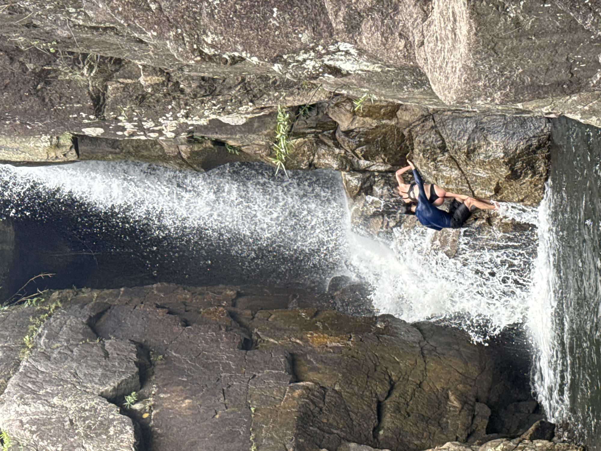 The image depicts a waterfall cascading over rocks in a natural setting. A person, dressed in a swimsuit with a dark long-sleeved shirt, is positioned on a rocky ledge near the waterfall's edge. The environment consists of rugged, dark-colored rocks and lush vegetation. The location is within the city of Đà Nẵng, Vietnam.