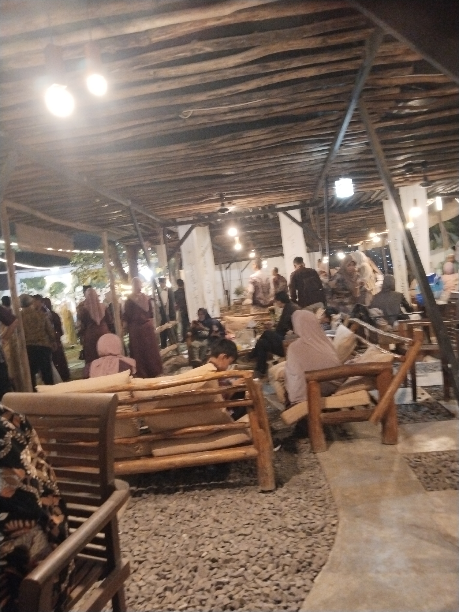 The image depicts an outdoor seating area with a rustic, natural theme, likely a cafe or restaurant, located in Serang, Indonesia. The ceiling is constructed from rough-hewn wooden beams, with exposed light bulbs illuminating the space. The flooring is a mix of gravel and concrete pathways. In the foreground, a person wearing a patterned garment is seated on a wooden bench. Scattered throughout the scene are other individuals, many of whom appear to be dressed in traditional Indonesian attire, including headscarves. People are engaged in various activities, such as conversing, sitting, and observing their surroundings. The overall ambiance suggests a casual and communal gathering space, possibly in the evening given the artificial lighting.