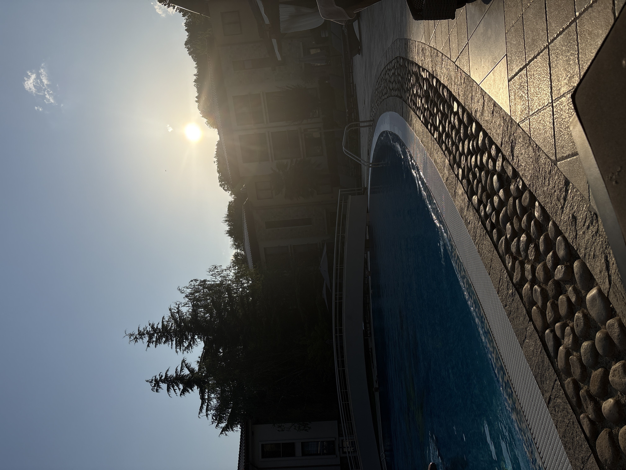The image depicts an outdoor swimming pool area during sunset. The sun is visible in the upper right quadrant of the sky, casting a warm glow over the scene. A large, blue swimming pool occupies the left side of the frame, with its edge defined by a white border and a decorative stone pathway. In the background, there are buildings and lush green trees on a hillside. A curved bridge spans a section of the pool. The paving stones around the pool are illuminated by the setting sun, creating a golden sheen. The overall impression is one of a serene and luxurious resort setting at the end of the day.