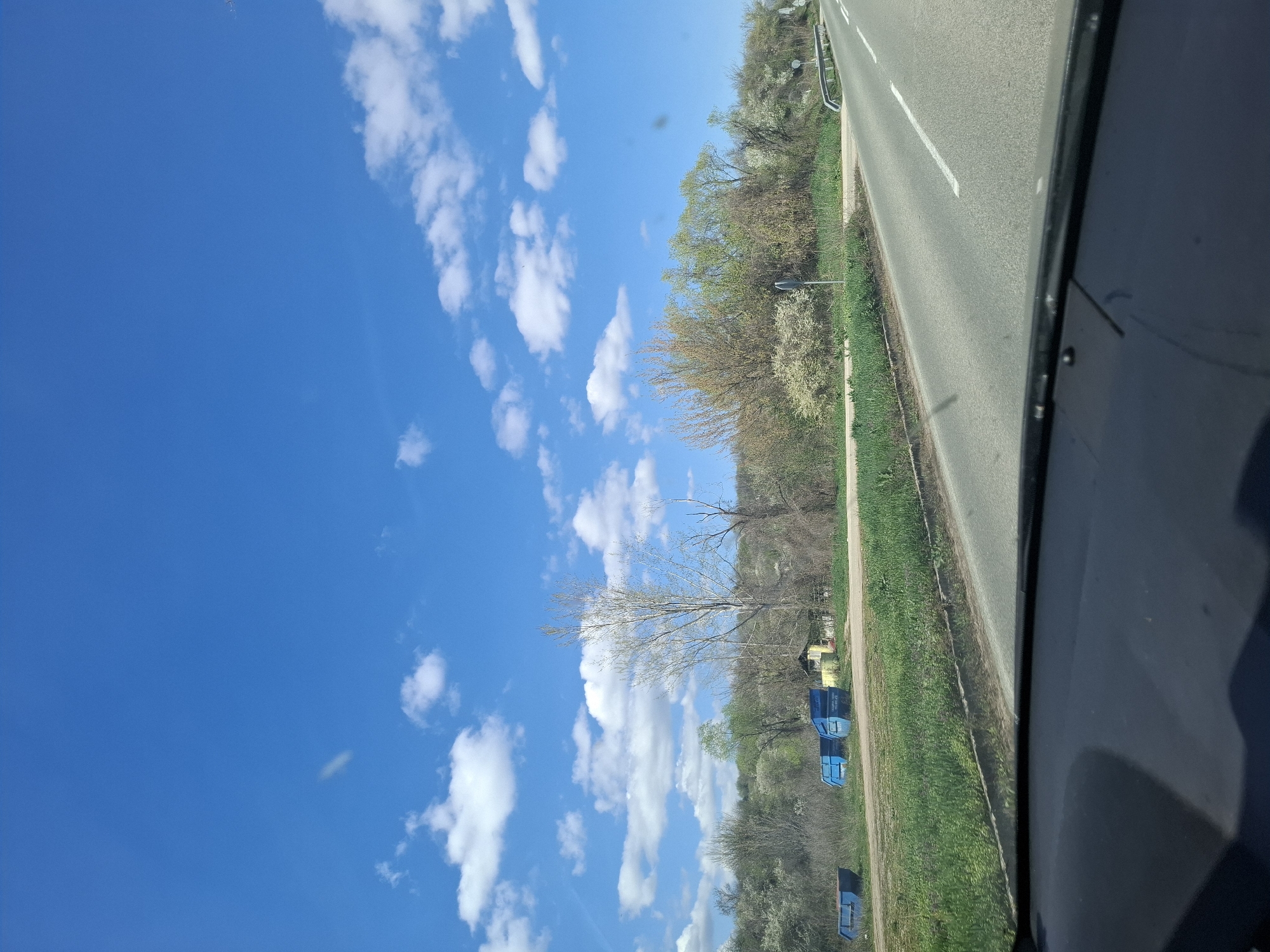 The image depicts a view from a vehicle, likely a car, traveling on a road in Tsar Simeonovo, Bulgaria. To the left is a blue sky with scattered cumulus clouds. To the right of the road is a grassy area and a line of trees. There are several blue metal containers or dumpsters visible along the grassy area. A two-lane road with a white dotted dividing line is adjacent to the grassy area. The edge of the car's windshield or window frame is visible on the right side of the frame.