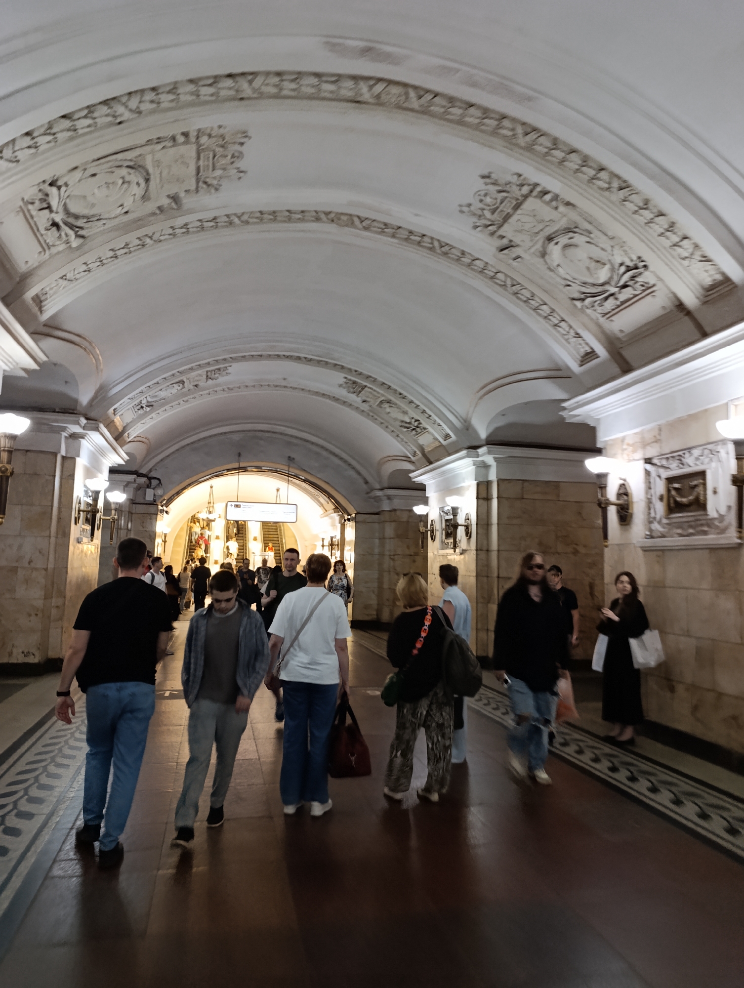 The image captures an underground pedestrian tunnel or subway station passage in Unknown, Unknown. The scene is illuminated by artificial lighting, showcasing a vaulted ceiling and tiled walls, with multiple people moving through the space.
The ceiling is a light-colored, arched structure, elaborately decorated with white plaster relief carvings. These carvings include repeating patterns of intertwined leaves or garlands, stylized geometric elements, and framed medallions positioned along the curves of the arch. The walls are clad in large, light-toned, possibly marble or stone, panels that exhibit subtle veining. Along the upper portion of the walls, beneath the ceiling, are decorative architectural moldings and wall-mounted sconces. Each sconce features multiple upward-facing light sources, casting a warm glow onto the ceiling and surrounding walls. The floor is a dark, highly reflective surface, possibly polished stone or wood, with a distinct lighter-colored border running parallel to the walls. This border displays a repeating dark pattern, likely a stylized leaf or scroll motif.
Approximately fifteen individuals are visible within the passage. Most are adults, dressed in casual clothing such as t-shirts, jeans, and light shirts. The majority of people are walking away from the camera, heading towards a brightly lit opening at the far end of the corridor where an escalator is ascending. In the immediate foreground, a man in a black t-shirt and blue jeans walks towards the left, while another man in a grey collared shirt over a t-shirt, dark pants, and sunglasses walks towards the center-right. A woman in a white t-shirt and blue jeans, carrying a dark brown shoulder bag, is seen walking to the right of the latter. Further back on the right, a man with long hair wearing sunglasses and a black top walks towards the camera. On the far right, a woman in a dark dress carries a white tote bag, also walking away from the camera. No significant interactions between the individuals are discernible; they appear to be primarily focused on their movement through the passage.
At the distant end of the tunnel, above the ascending escalator, a rectangular white sign with dark text and symbols is suspended. The escalator itself is visible, with several people being carried upwards. The overall lighting scheme provides consistent illumination throughout the passage, creating distinct reflections of the people and architectural elements on the polished floor. The environment appears clean and well-maintained, characteristic of a public transportation or pedestrian thoroughfare.