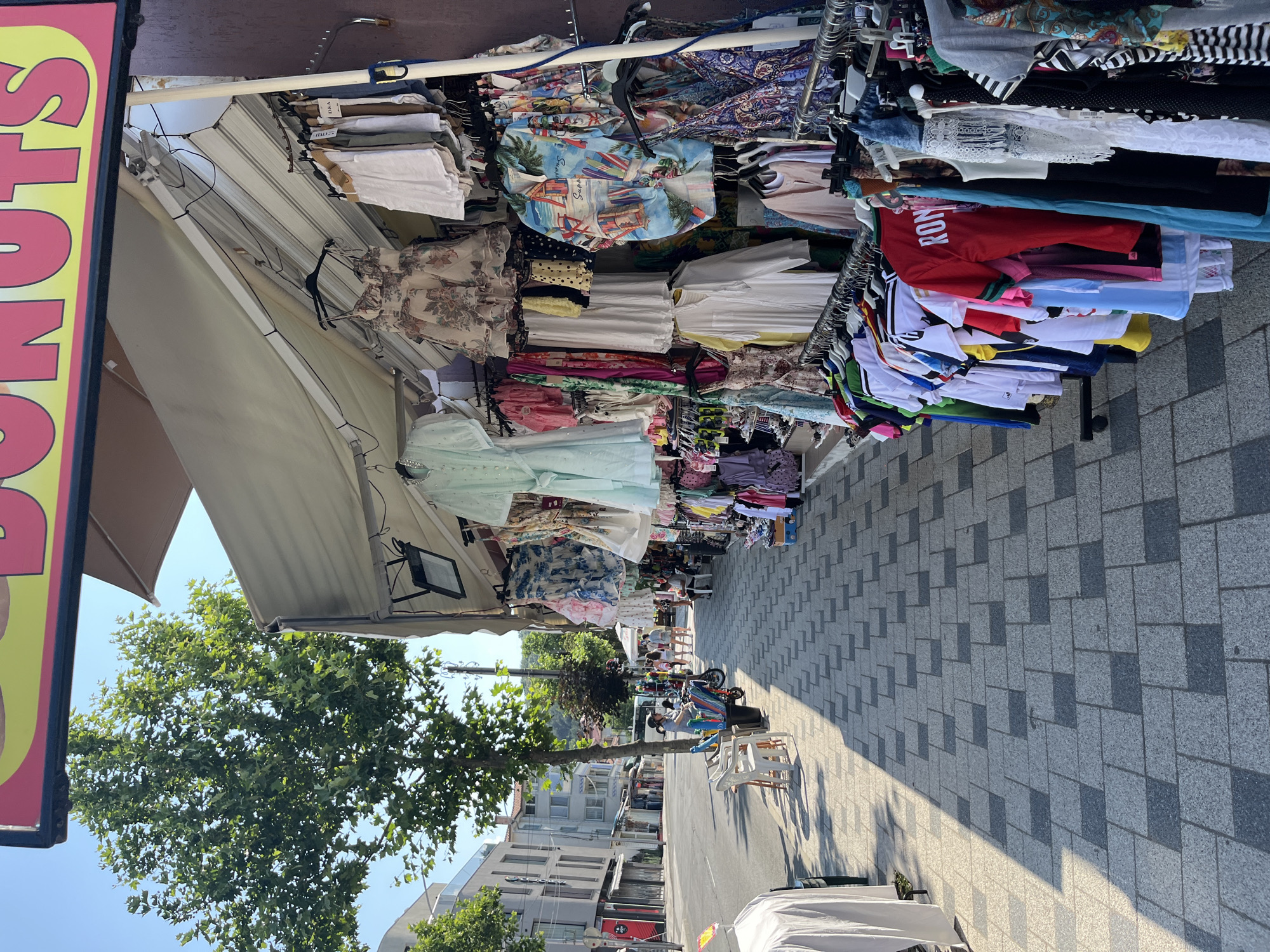 This is a tilted, eye-level shot of an outdoor clothing market stall in Obzor, Bulgaria. The scene is brightly lit, suggesting it is daytime, likely late morning or early afternoon, with clear blue skies and dappled sunlight casting shadows on the paved walkway. The stall is laden with a variety of clothing, primarily women's garments, displayed on racks and folded on tables. Items include colorful patterned tops, dresses, and what appear to be t-shirts and casual wear. The clothing is arranged in a somewhat haphazard but abundant manner.
In the background, a tree with lush green foliage is visible, along with buildings that appear to be part of a town or resort area. Several people are present, scattered along the walkway in front of and between stalls. Some are browsing, while others appear to be sitting or resting. One person is visible sitting on a white plastic chair in the foreground, facing away from the camera. The overall atmosphere suggests a lively and casual market environment, likely during a tourist season.
On the far left of the image, a portion of a large sign is visible. The text "DONUTS" is prominently displayed in large, yellow capital letters with a red outline. The sign is partially obscured by the angle of the shot and other elements of the market. The paving stones on the walkway feature a patterned design of light and dark grey rectangles.