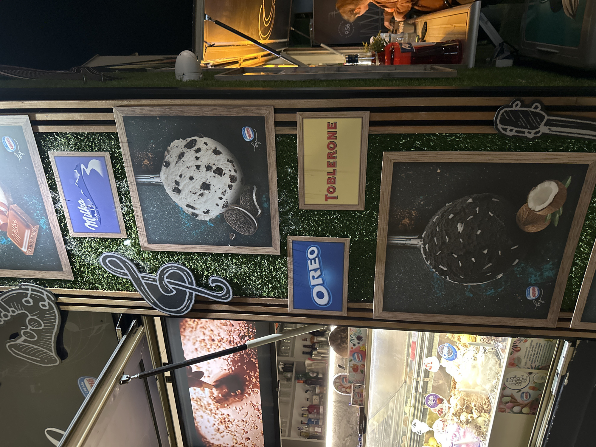 The image shows a stand in Veliko Tarnovo, Bulgaria, selling ice cream. Above the serving window are framed advertisements for ice cream flavors featuring Nestlé, Milka, Oreo, and Toblerone brands. The counter has an artificial grass surface decorated with music note cutouts. Behind the ice cream vendor, shelves of liquor bottles are visible. The setting is outdoors and illuminated by artificial lights.