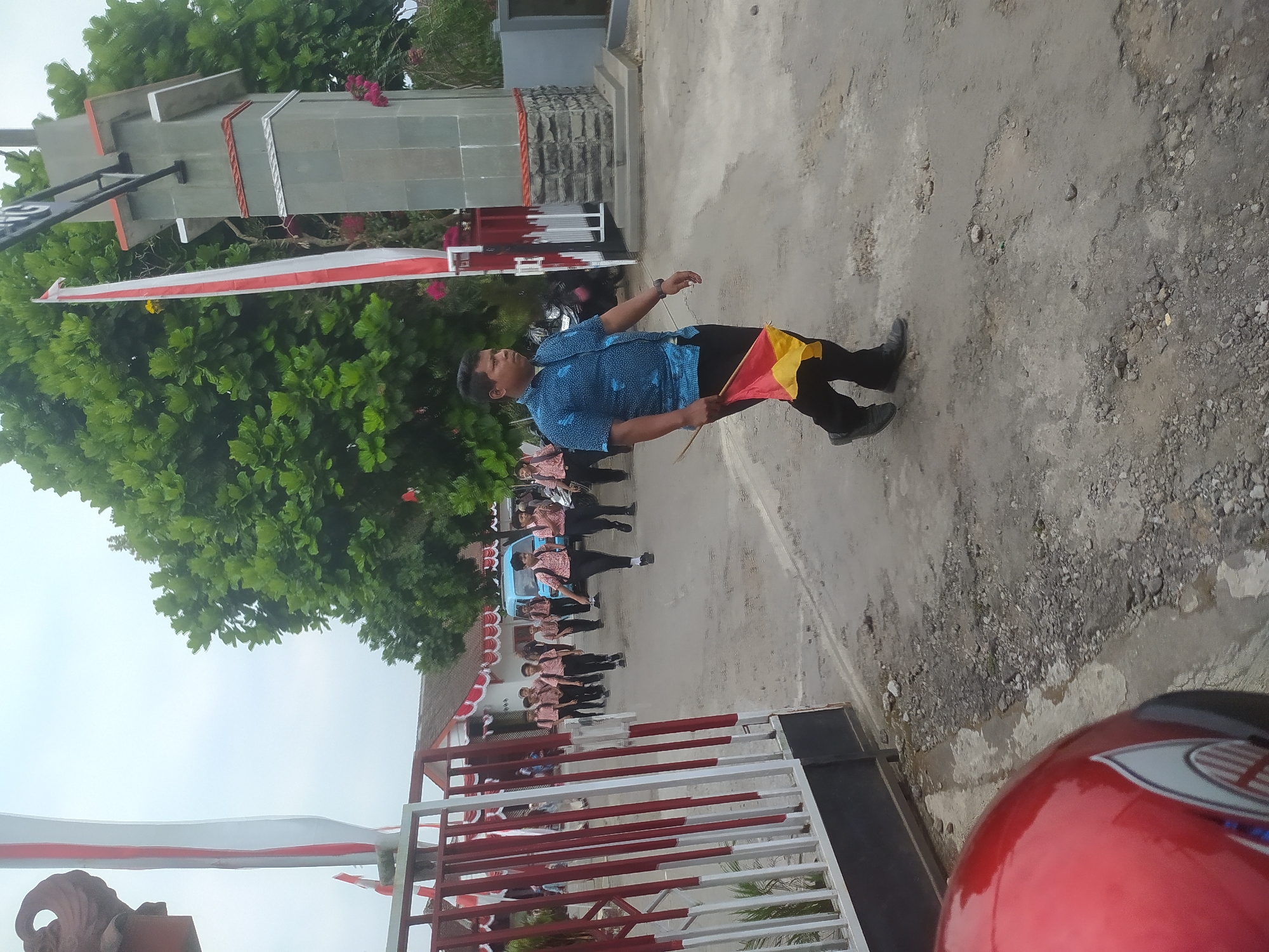 A man in a blue patterned shirt and black pants walks forward carrying a small red and yellow flag, on a dirt road in front of a school building in Candirejo, Indonesia. Behind him, a group of male students in uniform walk in the same direction, some carrying backpacks. A blue bus is visible parked on the road behind the students. The school building is decorated with red and white flags and banners. A large tree with green foliage is to the right of the image. A red motorcycle helmet is partially visible in the lower left corner.