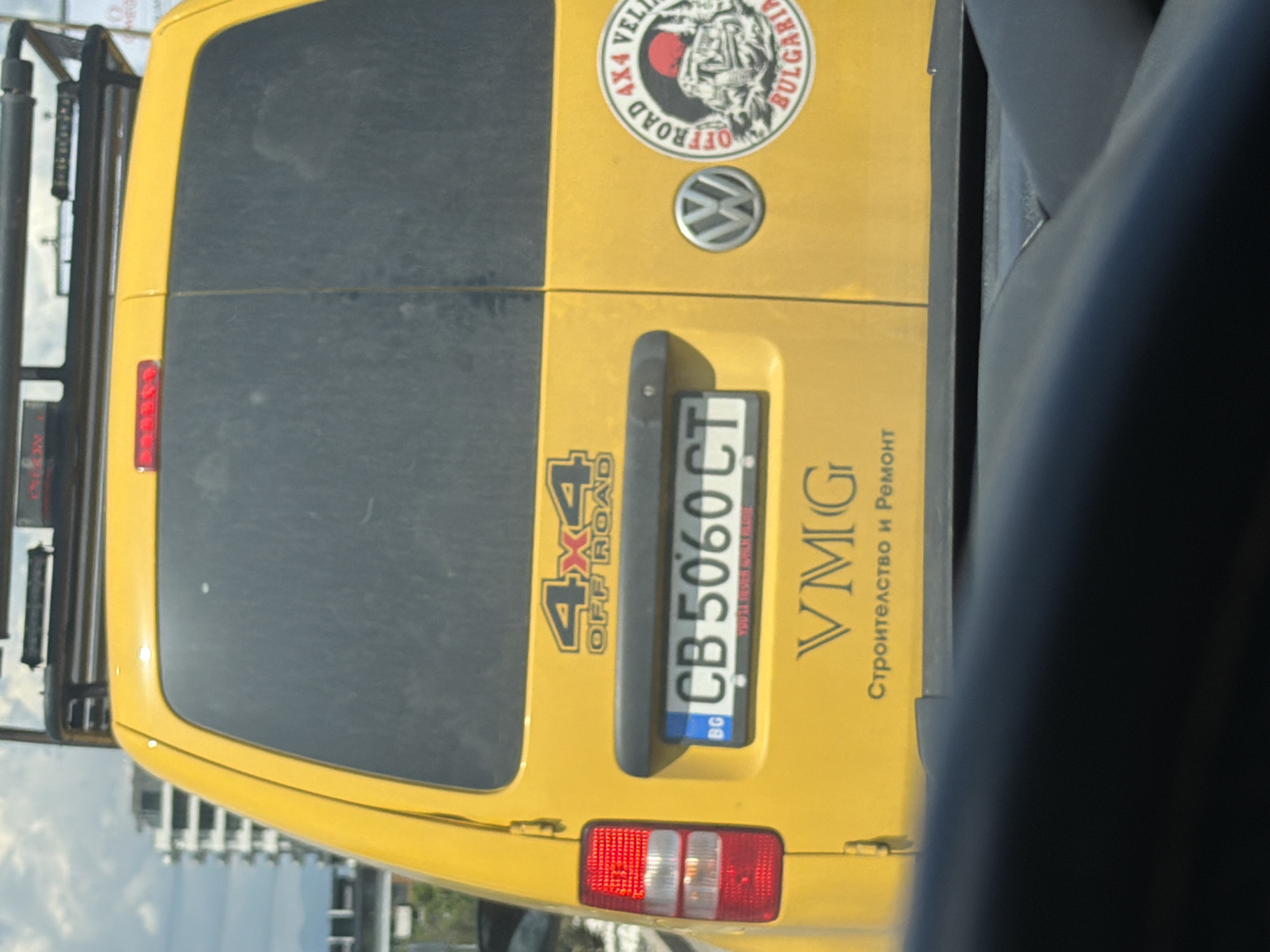 A yellow Volkswagen Caddy van, viewed from the rear, is parked in a city setting. The van has a roof rack and prominent stickers. One sticker, placed on the right side of the tailgate, reads "OFFROAD 4X4 VELIKO TARNOVO BULGARIA" and features an image of an off-road vehicle and a sun. Another sticker above the license plate says "4X4 OFF ROAD". The license plate reads "CB 5060 CT". Below the license plate, text in Cyrillic reads "VMG Строителство и Ремонт". In the background, a tall, modern building with many windows is visible. The sky is overcast with clouds. The van is situated on a street, likely in Sofia, Bulgaria, given the Cyrillic text and the context of Bulgarian license plates. No people are visible in the image. The scene appears to be captured during daylight.