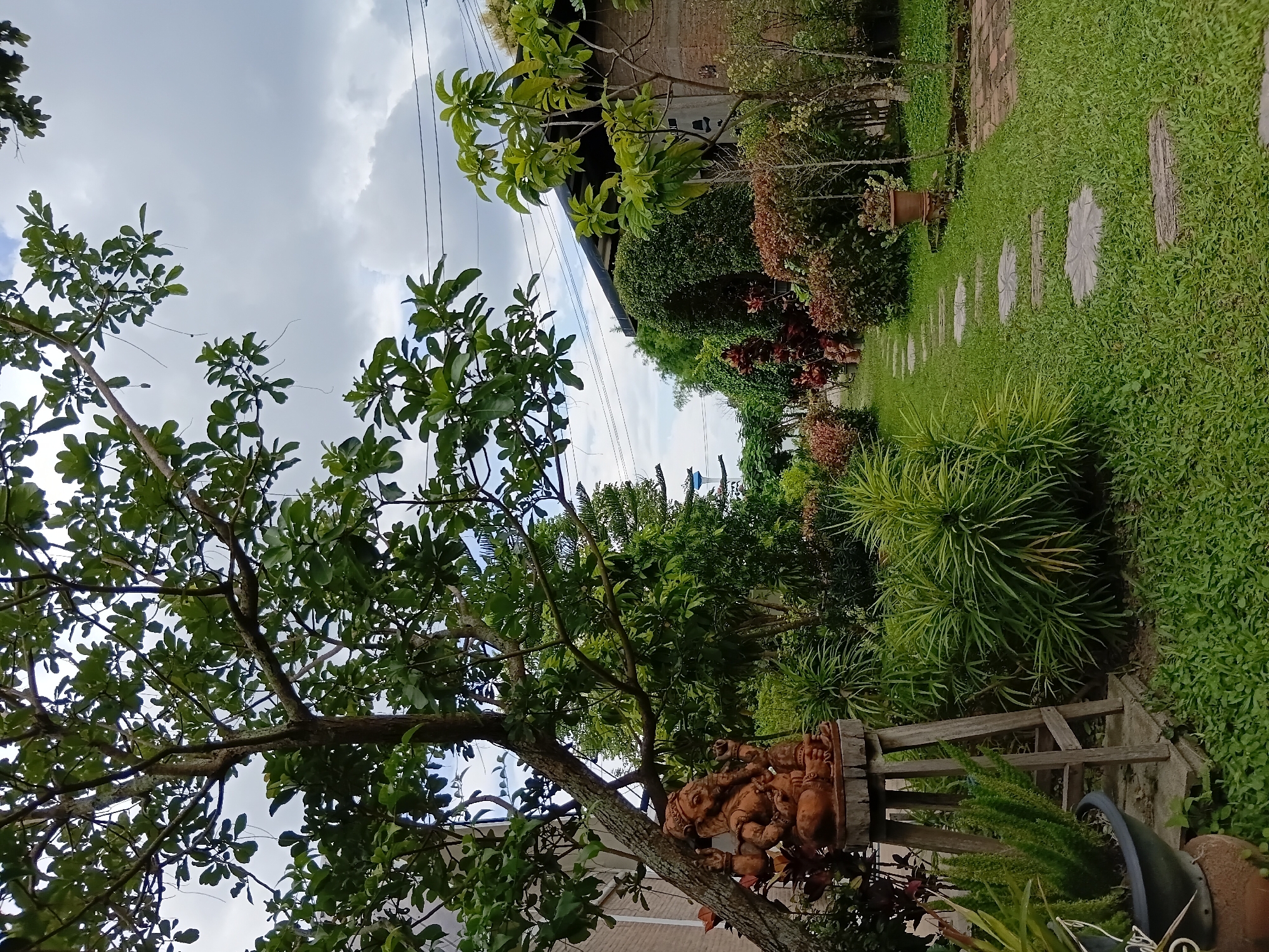 An outdoor scene depicts a garden with lush greenery, stone stepping stones, and a statue of the deity Ganesha. The garden is located in Ban Tong, Thailand. In the foreground, a brown terracotta statue of Ganesha is positioned on a wooden stand. To the left, a large tree with dark bark and green leaves provides shade. A paved walkway with circular stone pavers winds through the manicured lawn. Various plants and bushes with red and green foliage border the path. In the background, a brick building with a dark roof is visible, with power lines running overhead. The sky is overcast with white clouds. No people are present in the scene.