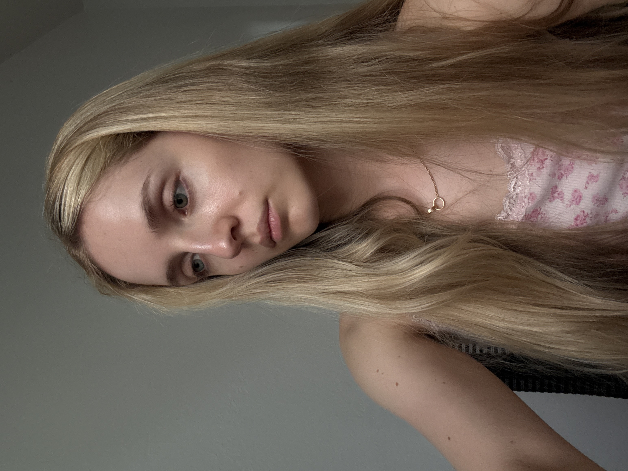 A young woman with long blonde hair is shown in a medium close-up, looking down and to the left. She is wearing a pink floral tank top with lace trim and a delicate gold necklace. Her skin appears fair, and she has light-colored eyes. The background is a plain, light grey wall. The scene is indoors, and there are no visible indicators of a specific location in Miami, United States of America.
