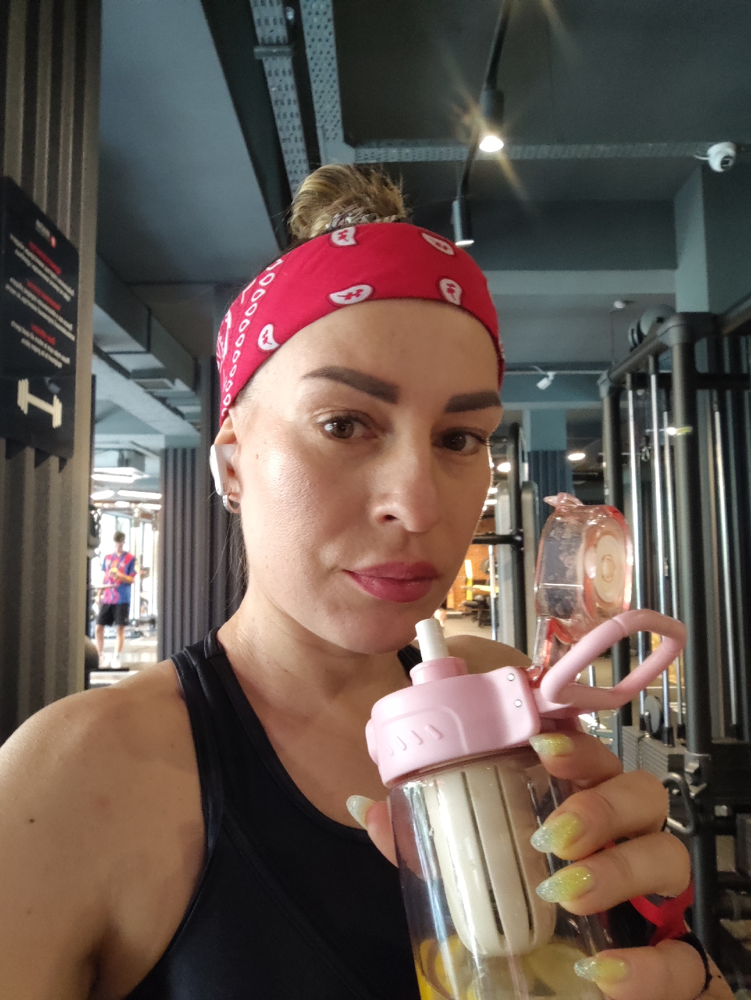 A woman is taking a selfie in a gym in Almaty, Kazakhstan. She is wearing a red bandana and a black sports bra. She is holding a pink water bottle with a straw. Her nails are painted yellow with glitter. In the background, another person is visible working out. The gym appears to be modern with metal equipment and track lighting.