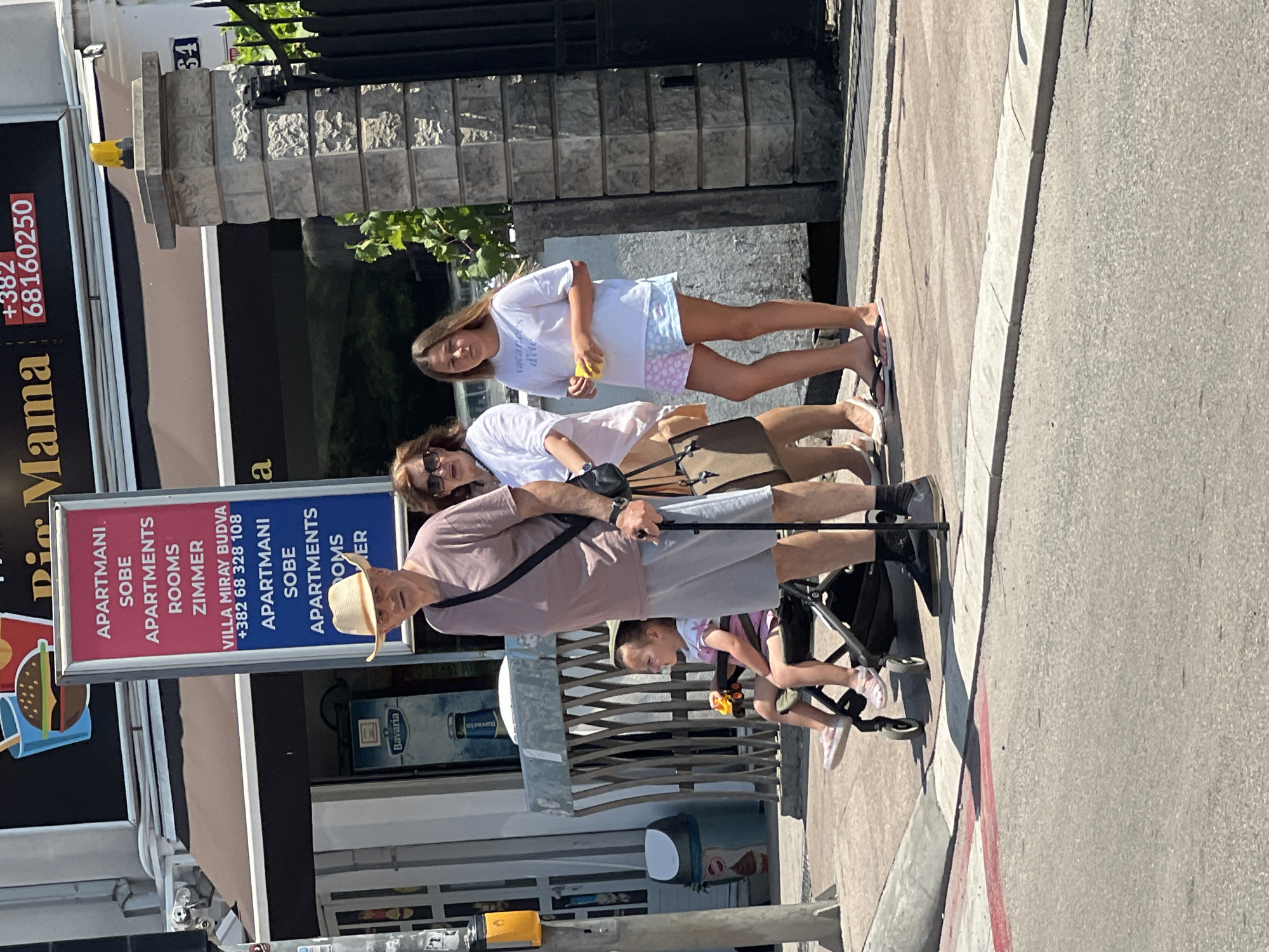 A group of four people, including two adults and two children, are standing on a sidewalk in Budva, Montenegro. The sidewalk is paved with light-colored concrete, and there is a crosswalk with red and white markings in the foreground. Behind the group, there is a building with a sign that reads "Big Mama" and "Apartmani Miray Budva." The building appears to be a hotel or apartment complex. A man in a straw hat and a pink shirt is using a cane. A young child is sitting in a stroller. A woman in a white shirt and shorts is standing next to the man. A young girl in a white t-shirt and patterned shorts is standing to the right of the woman. The weather is sunny.