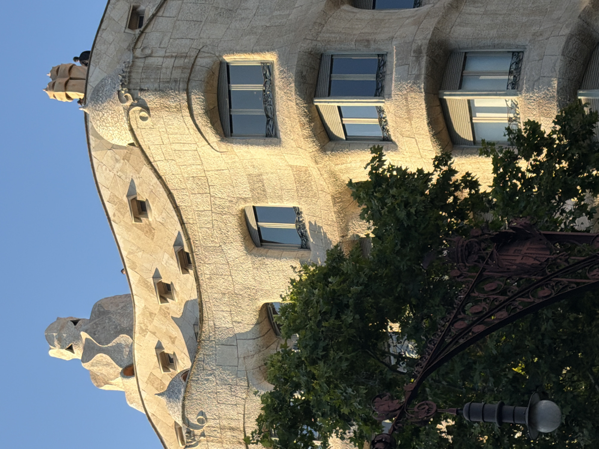The image shows the facade of Casa Batlló, a building located on Passeig de Gràcia in the Eixample district of Barcelona, Spain. The building's distinctive organic, undulating architecture, designed by Antoni Gaudí, is visible, characterized by its stone facade, curved windows, and decorative roof elements resembling a dragon's back. A large tree with green foliage partially obstructs the lower left of the building. In the upper portion of the image, the sky is clear and blue. Two people are visible on the roof of the building. A decorative lamppost is visible in the foreground on the left, made of dark metal with ornate scrollwork. The lighting suggests it is either early morning or late afternoon, with the sun casting long shadows and highlighting the textured surfaces of the building.