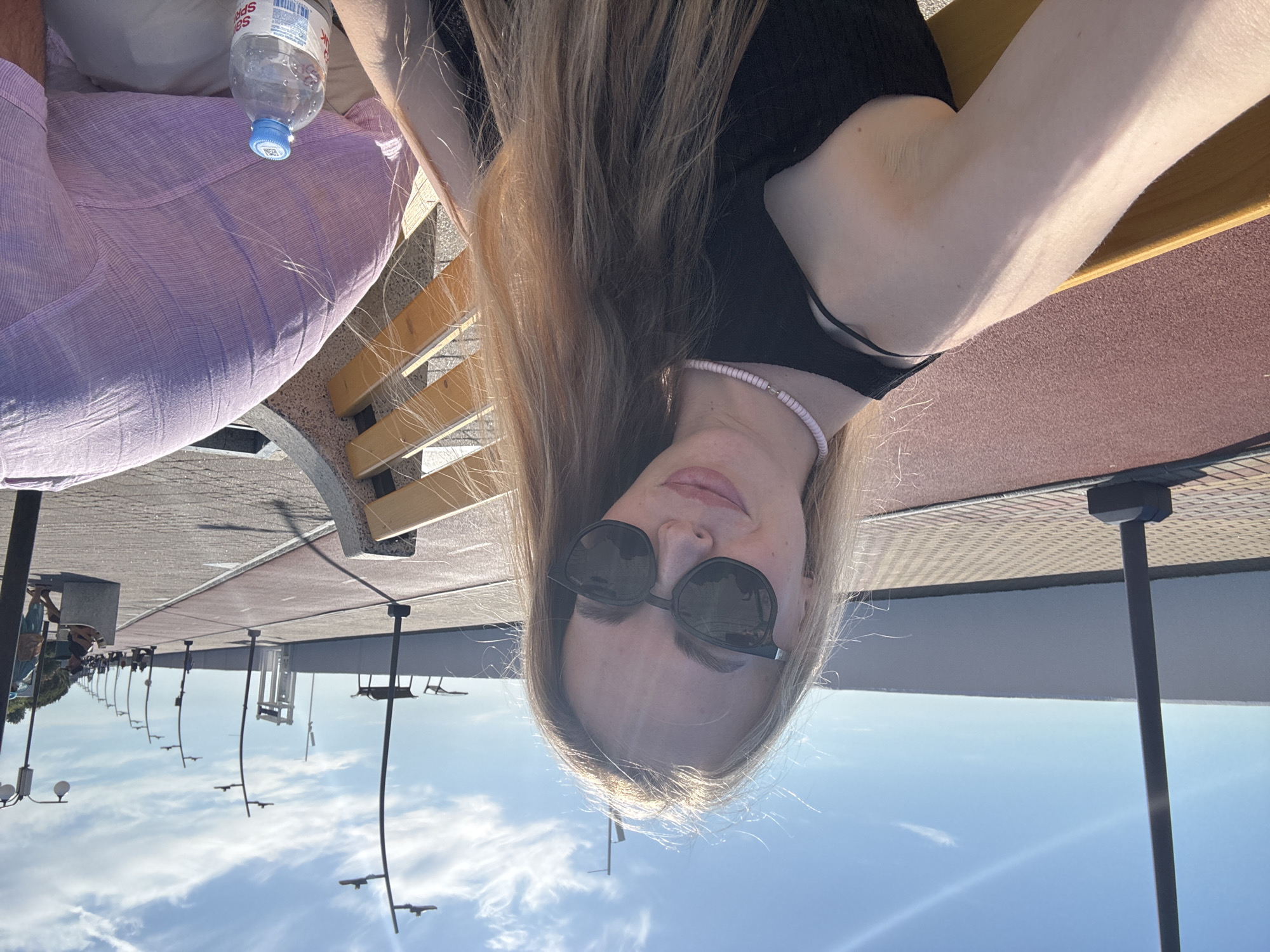A selfie or upward-angled shot captures a young woman with long, light brown hair, fair skin, and dark sunglasses, looking at the camera with a relaxed expression. She wears a black top and a light-colored beaded necklace. Sunlight highlights her hair and arm. She is seated, likely on a bench, with wooden slats visible to her right.
To her left, another individual is partially visible, wearing a light purple, possibly linen, shirt. A clear plastic Sprite water bottle, with its label and a blue cap, rests on their lap. Russian text ("Спрайт") is visible on the bottle.
The setting is an outdoor public area under a clear, bright blue sky with a few scattered white clouds. The ground features a paved walkway, with sections of grey concrete and a reddish-brown, textured surface that could be a running track or specialized paving. Numerous tall, dark streetlights or lampposts line the path, receding into the distance, suggesting a promenade or a modern park. The strong sunlight indicates it is daytime. The overall scene conveys a sense of leisure and relaxation in a sunny outdoor environment, consistent with a location like Sirius, Russia.