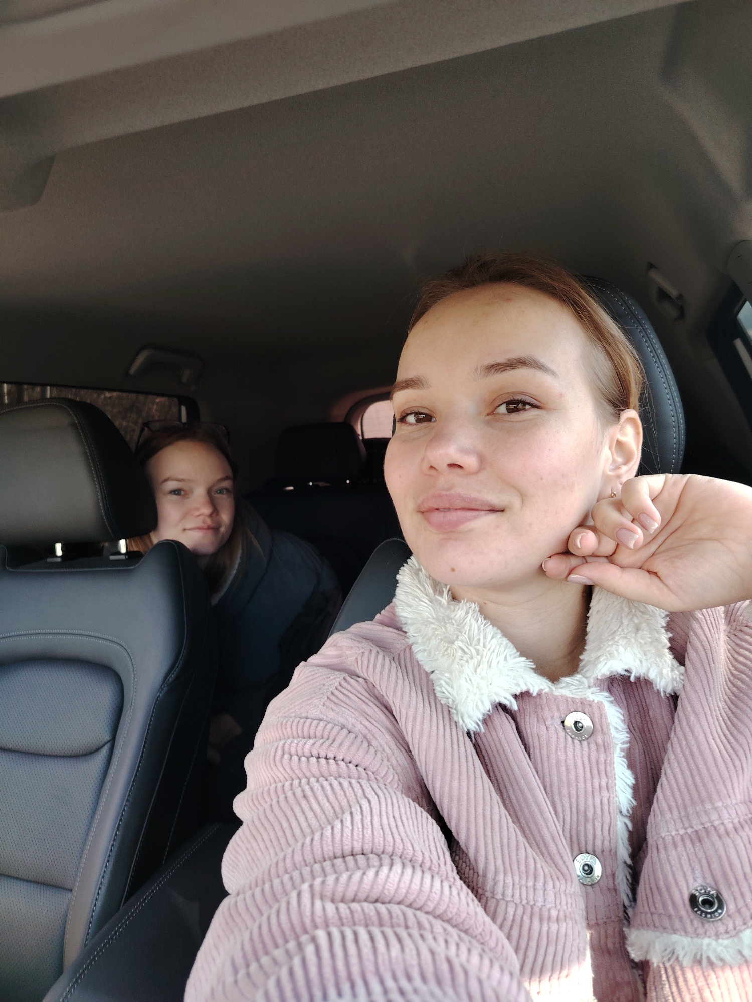 Two women are in a car, likely a selfie taken by the woman in the foreground. The woman in the foreground wears a pink corduroy jacket with a white fuzzy collar. She is resting her head on her hand. The woman in the background sits behind the driver's seat. The car interior includes black leather seats. The location is likely in Chaikovsky, Russia.