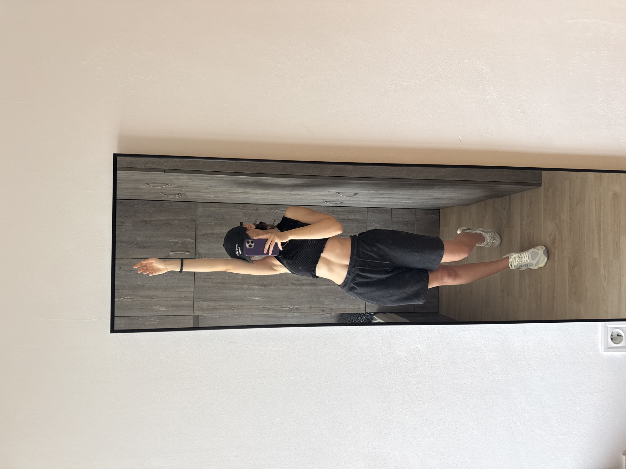 A young adult female takes a selfie in a mirror. She is wearing a black crop top, black shorts, a black baseball cap, and white sneakers. The background includes grey cabinets, a light wood floor, and a white wall with an electrical outlet. The location appears to be indoors, possibly an apartment or dressing room. No information within the image suggests a connection to Plovdiv, Bulgaria.