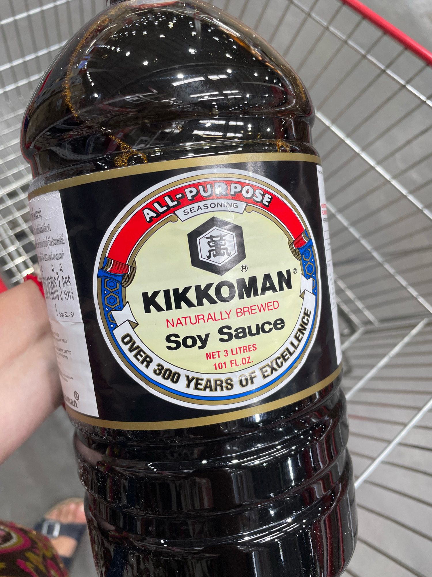 An extreme close-up, medium shot shows a large, clear plastic bottle of Kikkoman All-Purpose Soy Sauce, standing upright. The bottle is full of dark brown soy sauce, with some small air bubbles visible inside. The label is prominently displayed, featuring the Kikkoman logo, the words "ALL-PURPOSE SEASONING," "KIKKOMAN," and "NATURALLY BREWED SOY SAUCE." Below that, it states "NET 3 LITRES 101 FL.OZ." and "OVER 300 YEARS OF EXCELLENCE." A portion of a person's arm, wearing a patterned sleeve and a bracelet, is visible on the left side of the frame, holding the bottle. The background consists of the gray metal shelving of what appears to be a shopping cart. The scene is brightly lit, suggesting an indoor environment. The exact location in Pattaya City, Thailand, cannot be determined from the image alone.